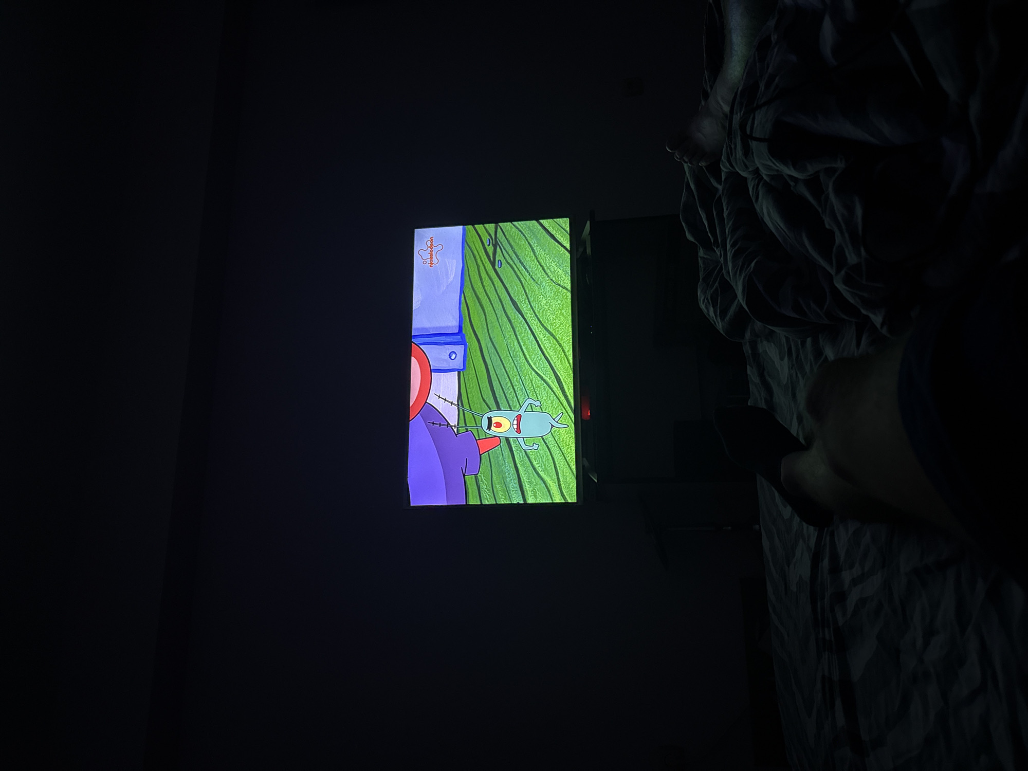 A dimly lit indoor scene shows a television screen displaying an episode of Spongebob Squarepants, featuring the character Plankton. A person is present, holding a remote control. The scene includes a bed. The location cannot be determined to be Burgas, Bulgaria based on the visual information provided.