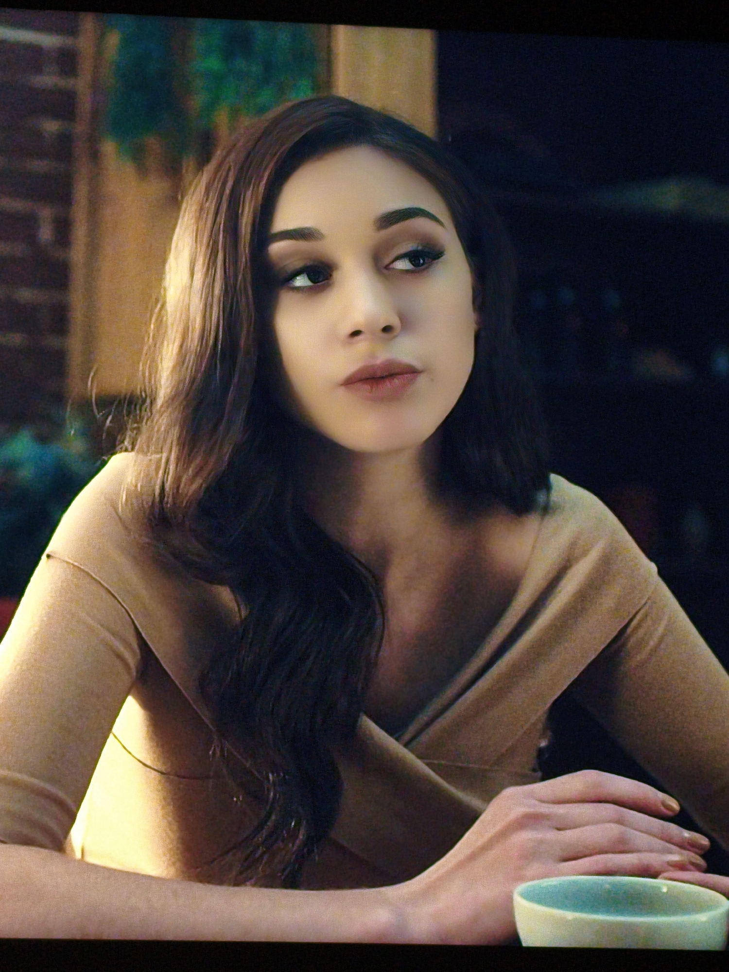 The image shows a close-up, head-and-shoulders shot of a young woman with long, dark, wavy hair, and light skin. She is wearing a tan, off-the-shoulder top. Her left arm is resting on a surface, and her hands are clasped together in front of her. To her right, there is a small, light blue and white bowl. The background is dark and appears to be an interior setting, possibly a restaurant or home, with hints of brickwork and foliage. There is no visible indication of the city of Vladivostok, Russia in the image.