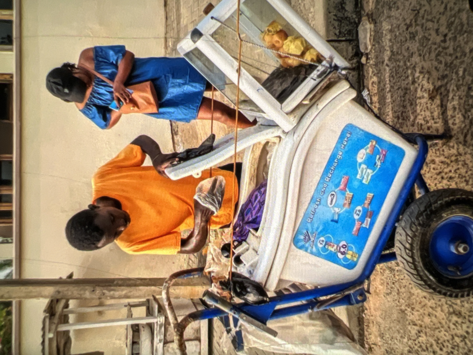 A young Black man in an orange t-shirt is standing behind a blue and white pushcart, handling a plastic bag. To his right, a Black woman wearing a blue off-the-shoulder dress and a black baseball cap is holding a small brown purse and a mobile phone. The pushcart contains a white cooler with a blue lid, adorned with a sticker depicting a cycle of charging devices. A transparent container with a metallic frame, filled with yellow items, is attached to the side of the cooler. The scene appears to be a street in Lagos, Nigeria, with a light-colored wall and a building with windows visible in the background. The ground is a mixture of sand and small stones.