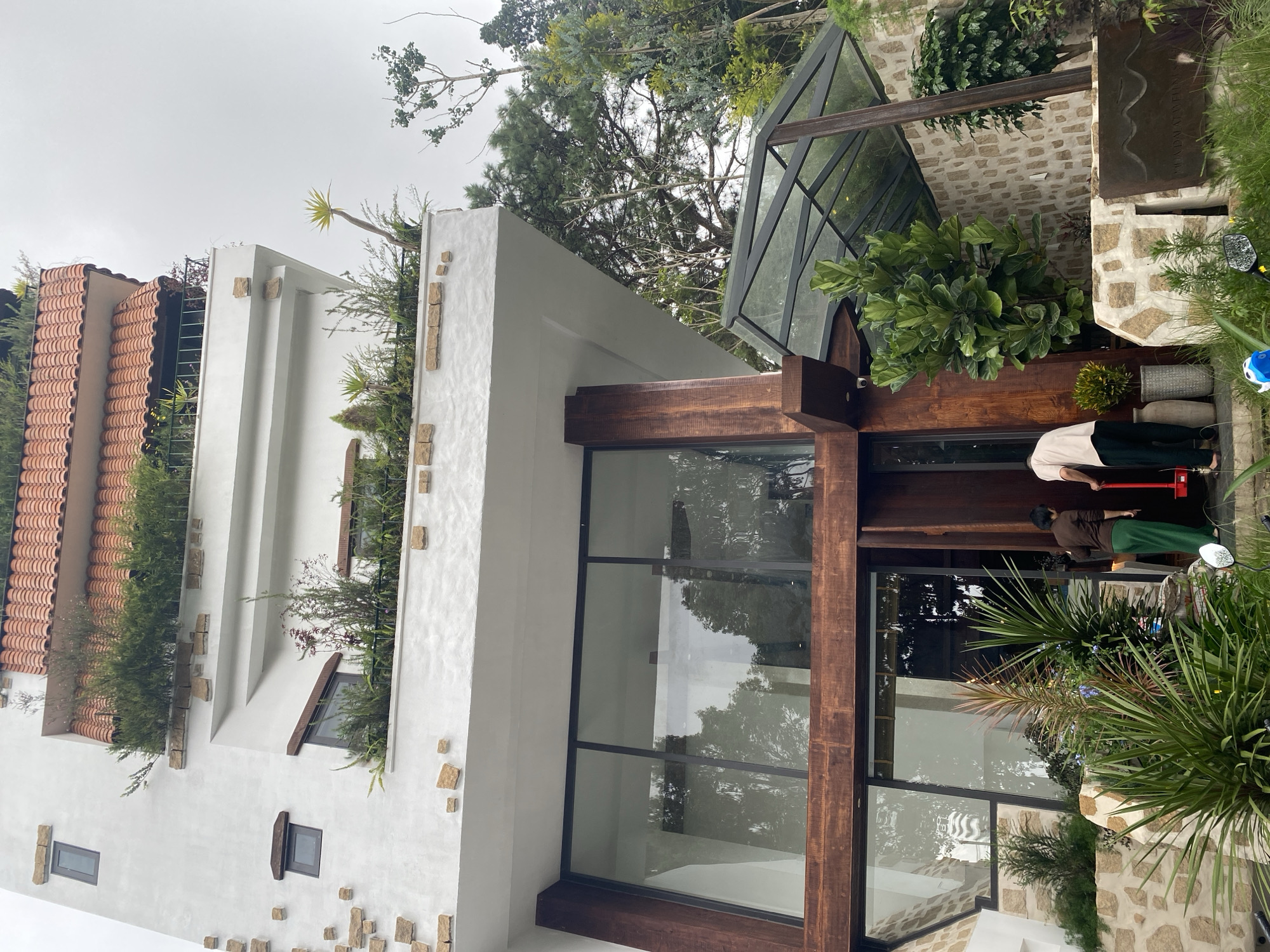 This image depicts a multi-story building with a unique architectural style, located in the Lâm Viên - Đà Lạt region of Vietnam. The building features a white stucco exterior with decorative stone accents and multiple balconies filled with lush greenery and plants, topped with a red tiled roof. The ground level has large glass windows and wooden structural elements. Two individuals are visible near the entrance; one is wearing a dark brown top and green pants, and the other is wearing a light-colored shirt and dark pants. They appear to be interacting with a red object, possibly a cleaning tool, near an open doorway. The surrounding environment includes various trees, plants, and stone landscaping, suggesting a natural and possibly resort-like setting. The sky is overcast, indicating a cloudy day.