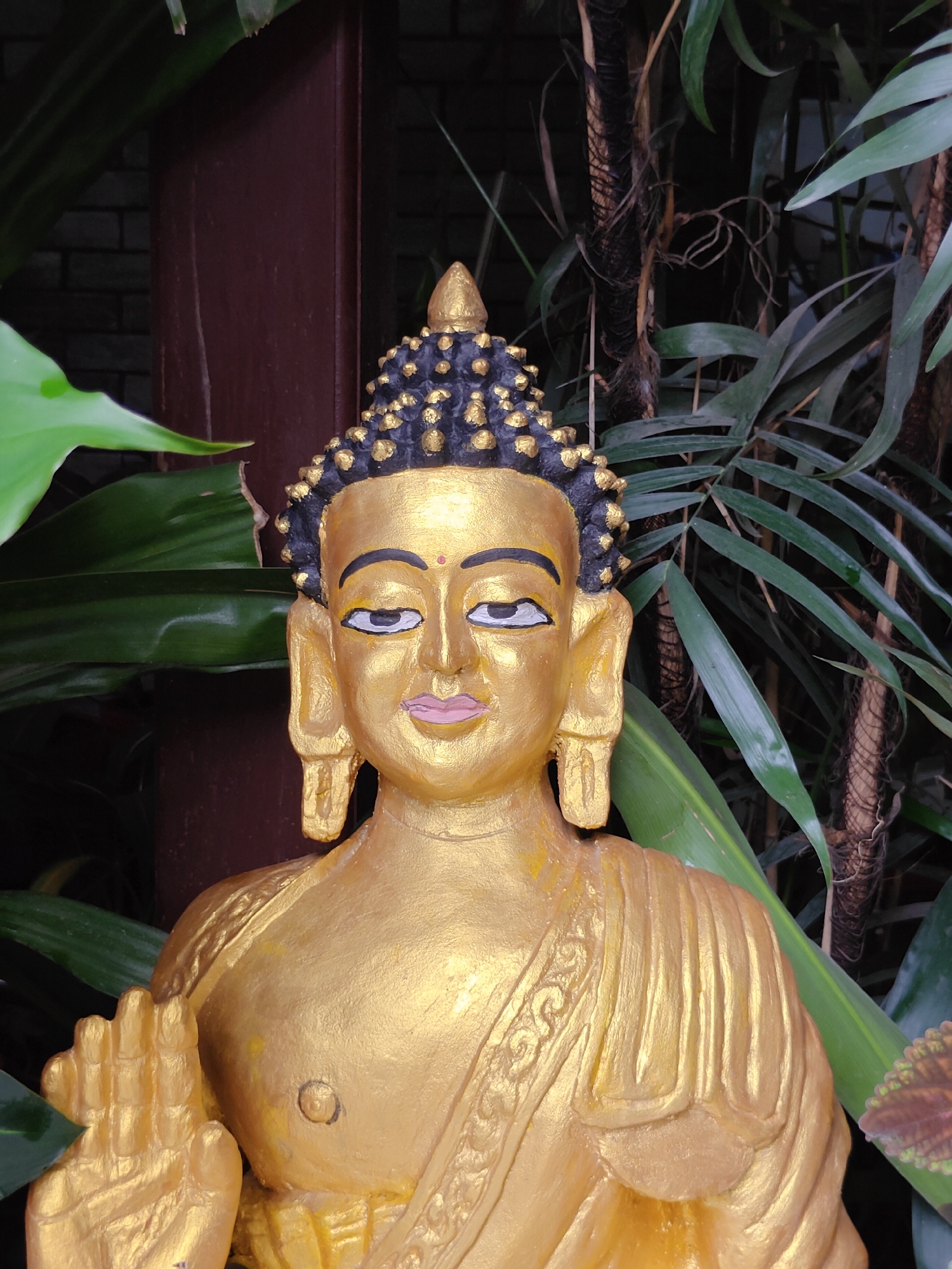 A golden Buddha statue is positioned in the foreground, facing forward. The statue is detailed with dark, curly hair on its head, painted eyes with black outlines, and pink lips. Its right hand is raised, palm facing outward, in a gesture of blessing or protection. The statue wears a robe draped over its left shoulder, adorned with a decorative pattern. Behind the statue, lush green foliage and tall, dark stalks create a natural, somewhat overgrown environment. To the left of the statue, a dark brown, vertical wooden pillar is visible, and in the background, a dark brick wall can be discerned. The image appears to be taken outdoors, possibly in a garden or a courtyard in Pokhara, Nepal, given the presence of tropical-looking plants and the cultural context of Buddha statues.