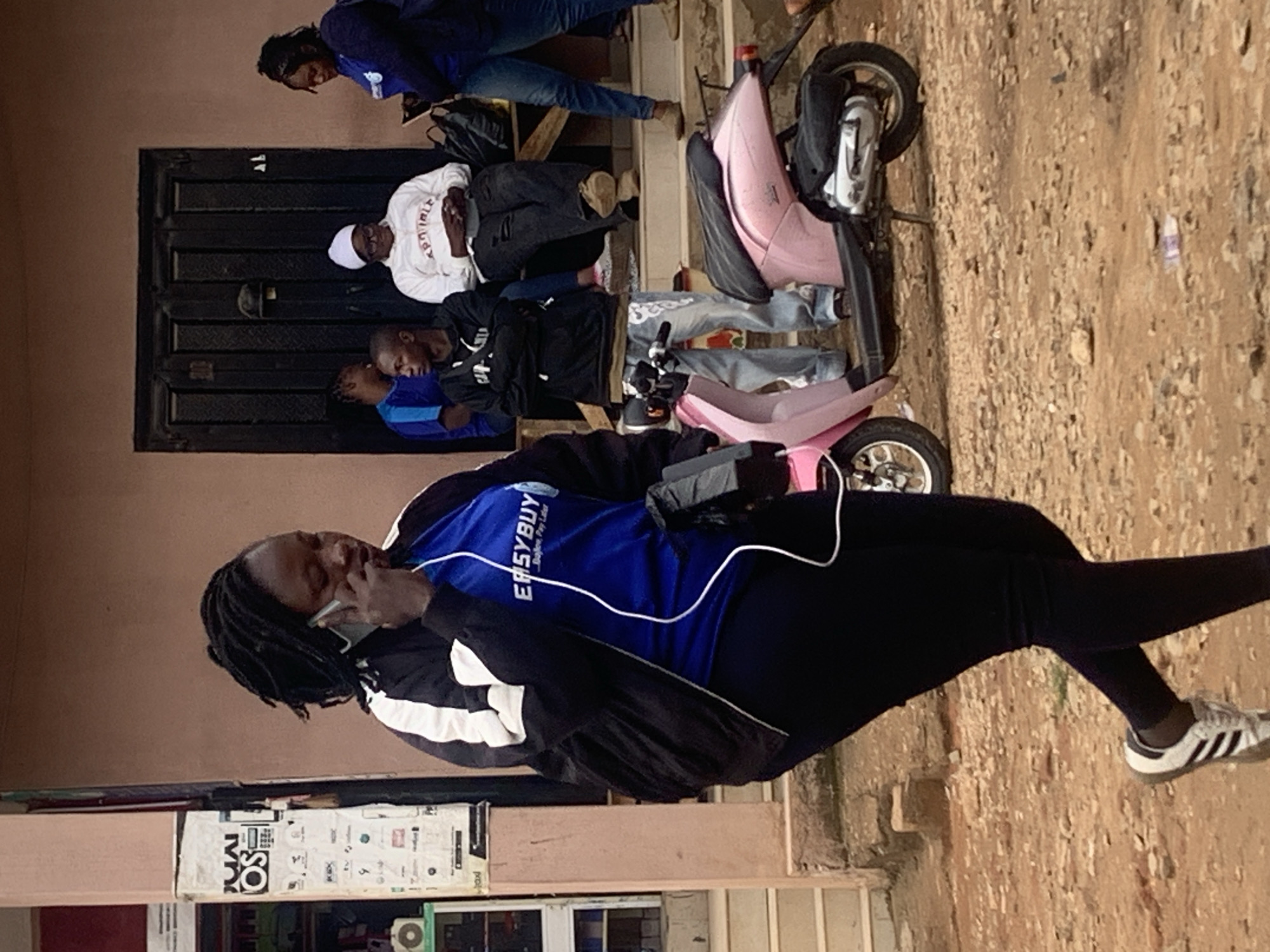 This image depicts a street scene in Jos, Nigeria. In the foreground, an African woman with dark skin and dreadlocks walks from left to right. She is wearing a black and white track jacket over a blue t-shirt featuring the white "EASYBUY - Buy Now, Pay Later" logo, black leggings, and white sneakers with black stripes. She holds a silver-colored smartphone to her right ear with her right hand, with white earbud wires extending downwards. A small black pouch or device with a white wire is attached to her left hip.
In the background, against a light brown building with a dark, vertically paneled double door, a group of four individuals is situated on a wooden bench or ledge. On the far left, partially visible, is a person in a blue shirt. Next, a young man with dark skin wears a black hoodie with white text and light blue patterned jeans, seated with a dark strap across his chest. Centrally, an older individual with dark skin wears a white beanie, glasses, and a white hooded sweatshirt with red text, seated with hands clasped. On the right, a woman in a dark blue long-sleeved shirt and light blue jeans stands or sits, holding a mobile device. Black bags are visible on the ground near the seated individuals.
A pink moped/scooter is parked on its kickstand in the midground, positioned between the walking woman and the background group. To the far left, a light-colored building pillar is extensively covered with white paper advertisements and stickers, some inverted. A small green cabinet with a circular opening is located at the base of this pillar. Light-colored steps lead up to the building's entrance. The ground is an uneven, reddish-brown dirt and gravel surface with scattered small stones. The scene is illuminated by natural daylight.