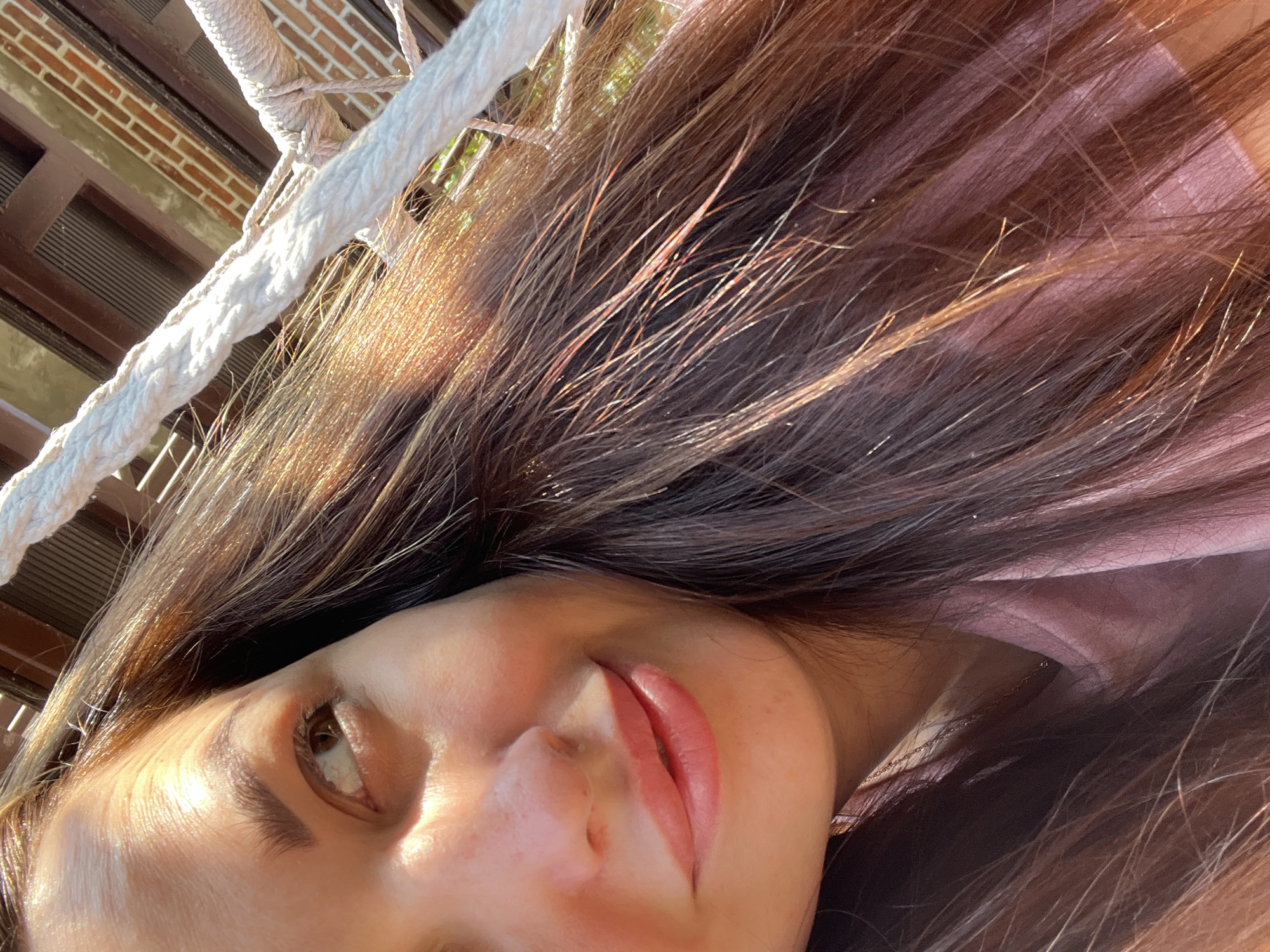 A close-up, over-the-shoulder shot captures a woman's face and hair. She is looking to her left, with the sunlight illuminating her forehead and cheek. Her lips are slightly parted, and she has a gentle smile. Her long, brown hair is styled down, with strands catching the light. She is wearing a pink t-shirt, and a white rope runs diagonally across the image, suggesting she might be in a hammock or on a boat. The background shows a brick wall and wooden beams, hinting at an outdoor setting. The overall impression is one of a relaxed and pleasant moment.