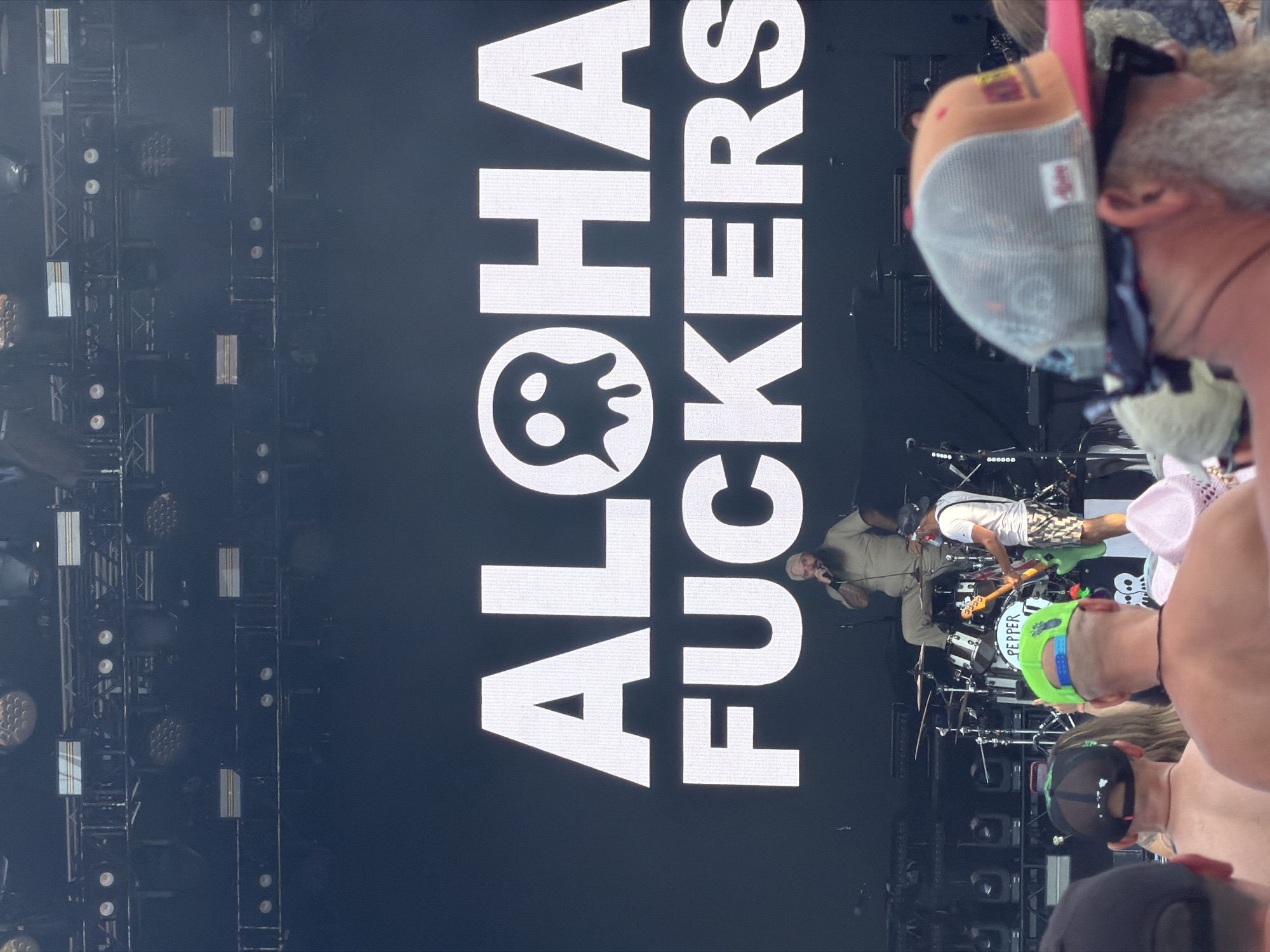 This image captures a concert scene at the Veterans United Home Loans Amphitheater in Virginia Beach, Virginia. A large, bright white LED screen displays the words "ALOHA FUCKERS" in prominent block letters, with a stylized ghost-like icon replacing the 'O' in "ALOHA." In front of the screen, on stage, a man with a beard is seated on what appears to be a drum stool and is speaking into a microphone. To his right, a musician is standing and playing a green electric guitar. Behind the seated man, a drum set is visible. The foreground of the image shows the backs of several concert-goers, many wearing baseball caps, indicating an outdoor event. The lighting suggests it is either late afternoon or evening.
