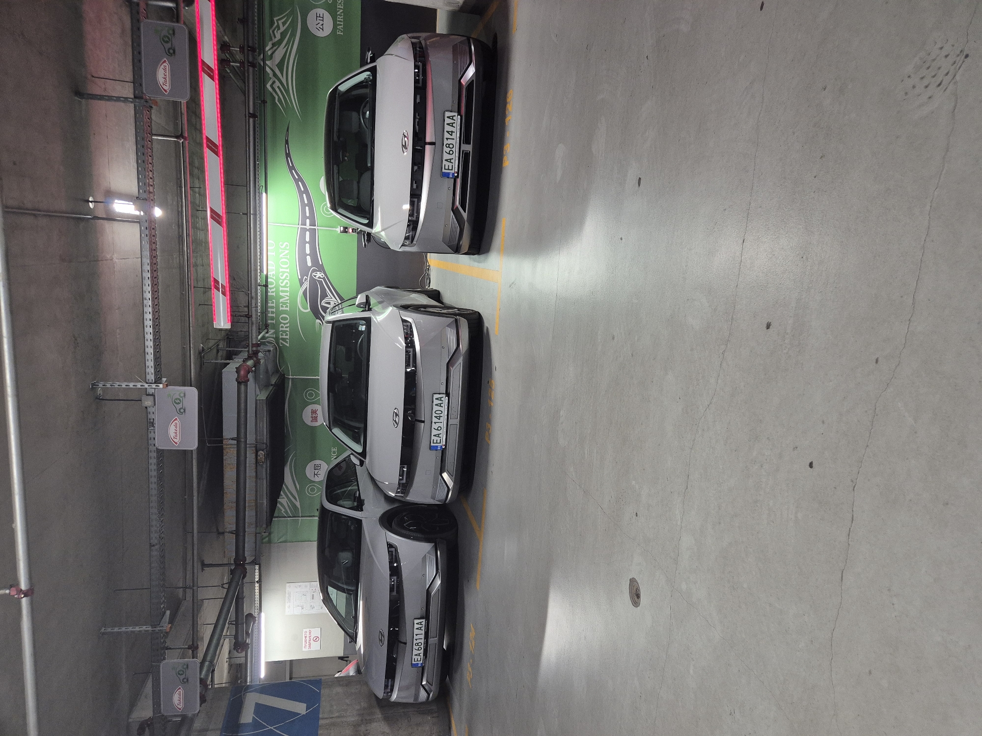 The image captures three silver Hyundai Ioniq 5 electric vehicles parked in a multi-story parking garage. The vehicles are aligned with their front bumpers facing the camera. The parking garage features a concrete floor with yellow painted parking lines. Overhead, there are exposed pipes, electrical conduits, and lighting fixtures. Two signs with the logo "Takeda" are visible hanging from the ceiling, and a large green banner with a stylized road and mountains, along with the text "ON THE ROAD TO ZERO EMISSIONS" and some Asian characters, is mounted on the wall behind the cars. Red and white striped barriers are also present overhead. The scene appears to be well-lit by artificial light, typical of an indoor parking facility. The license plates of the cars are visible, with the leftmost reading "EA 6811 AA", the middle one "EA 6140 AA", and the rightmost "EA 6814 AA". A circular drain is present on the concrete floor in the foreground. The overall environment suggests a modern parking facility, likely equipped for electric vehicle charging given the banner. Based on the license plate format (EA followed by numbers and letters), this location is likely in Bulgaria, and considering the context of "Takeda" and "Zero Emissions" branding, it could be a corporate parking lot or a public charging station in Sofia, Bulgaria.