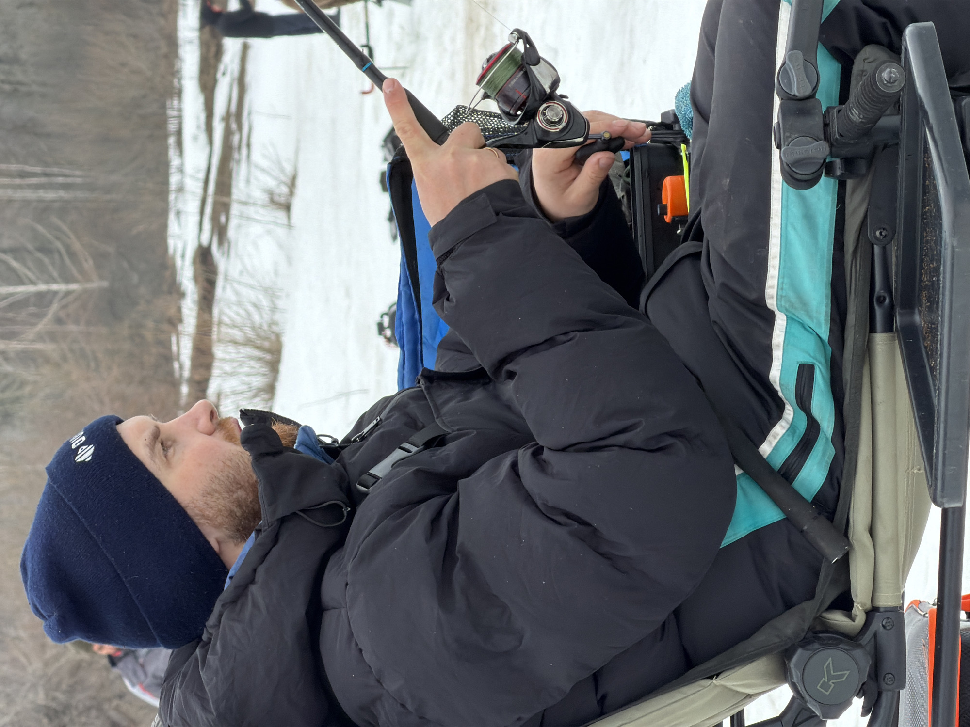 The image depicts a man ice fishing in Noviy, Russia. He wears a dark blue beanie, a black winter jacket, and gloves. He is holding a fishing rod and reel, facing the ice-covered terrain. The man sits in a chair attached to a wagon, which appears to be positioned on the frozen surface. A white, snowy landscape extends into the background, with indications of trees. The man appears to be manipulating the reel of his fishing rod. The wagon's frame is visible, with straps, fabric, and other equipment attached.
