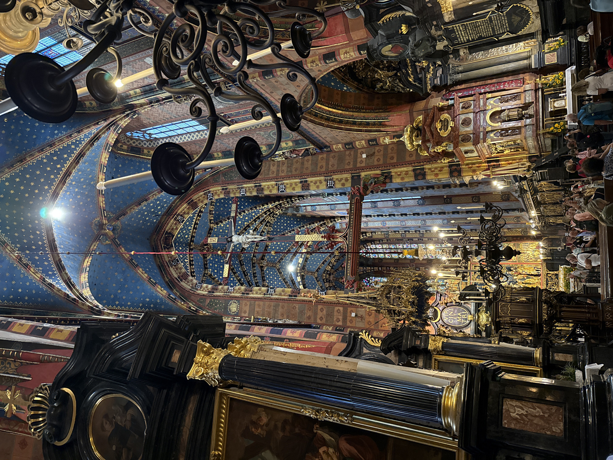 The image shows the interior of St. Mary's Basilica in Krakow, Poland. The basilica features a high, vaulted ceiling adorned with a star-like pattern in shades of blue and gold. Elaborate chandeliers hang from the ceiling, casting light on the ornate architecture. The walls are decorated with colorful patterns and religious imagery, including a prominent crucifix with Christ's figure. In the foreground, the richly decorated interior includes a large pillar with gilded details and framed artwork. A group of people are visible in the lower portion of the image, standing and observing the basilica's interior. Some are taking photographs. The overall impression is one of grandeur and religious significance.