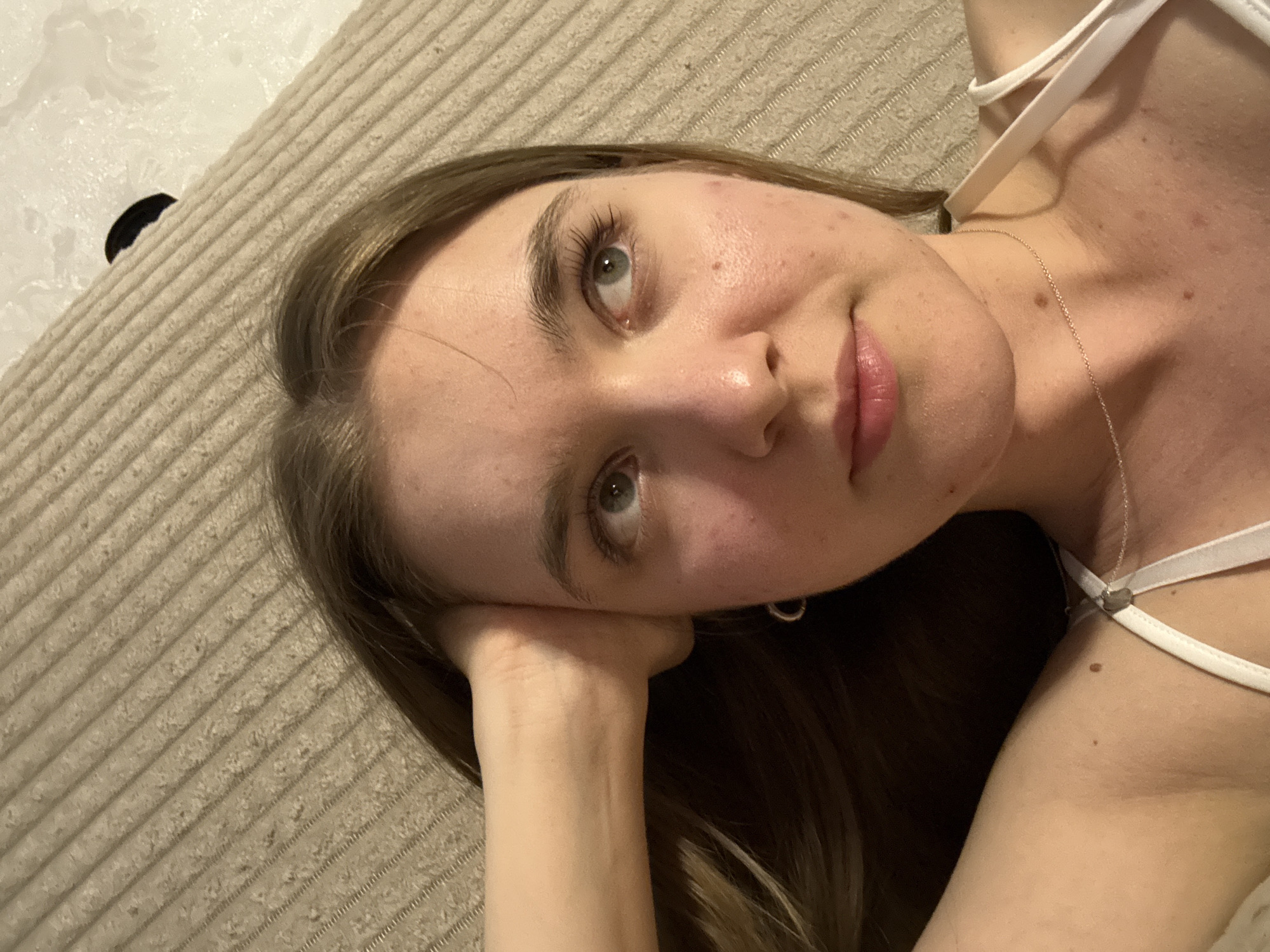 A fair-skinned woman with light brown hair is lying on a beige corduroy surface. Her head rests on her left hand. She wears a white top with thin straps and a silver necklace. Her gaze is directed towards the camera. A black object is partially visible in the upper left corner of the image. The location is unspecified.