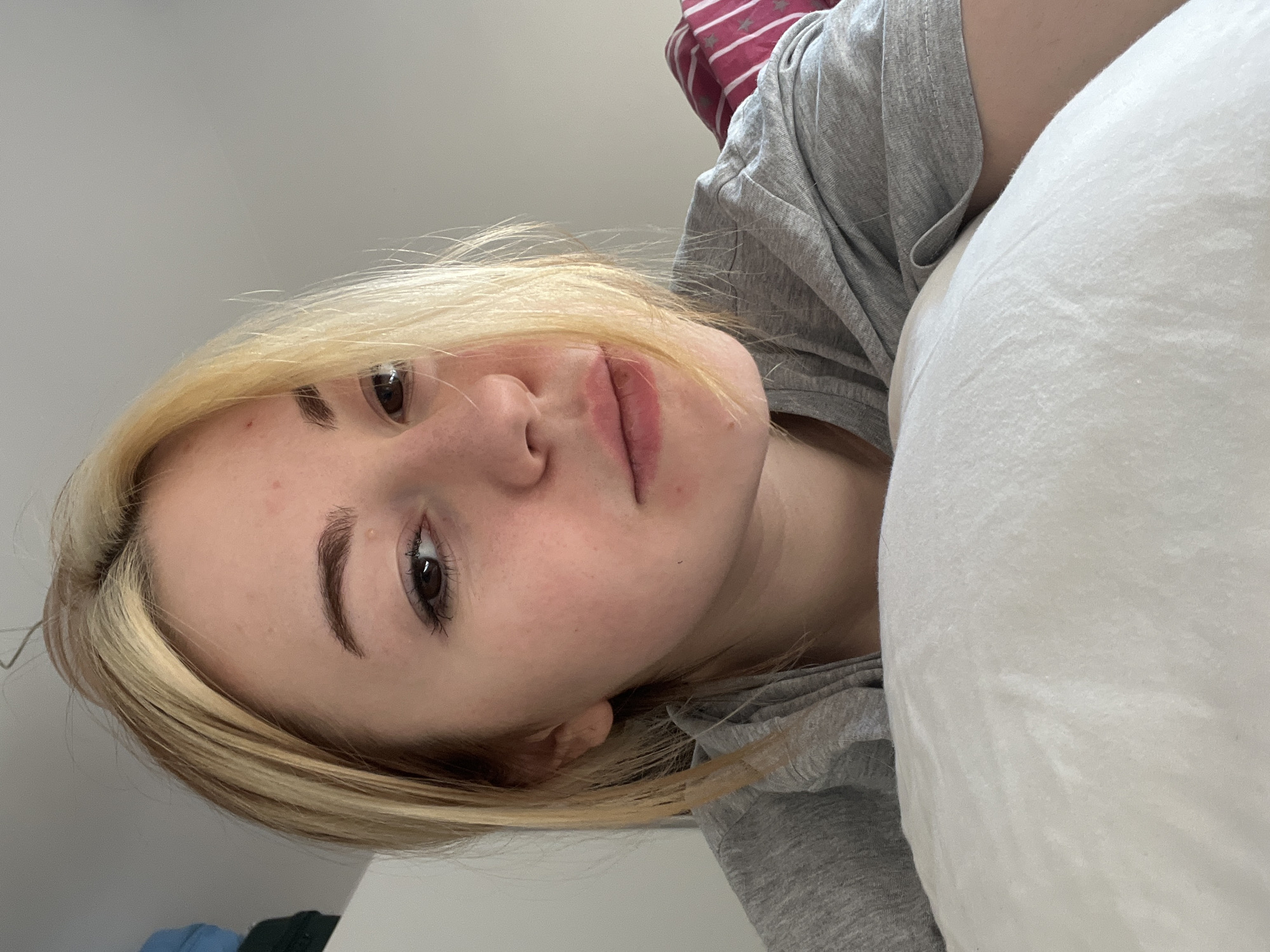 A close-up, upside-down selfie of a young woman with light blonde hair and fair skin. She is wearing a gray t-shirt and appears to be lying on a white pillow. The background is a plain white wall. The location in the city of Boreti, Montenegro cannot be determined from this image.