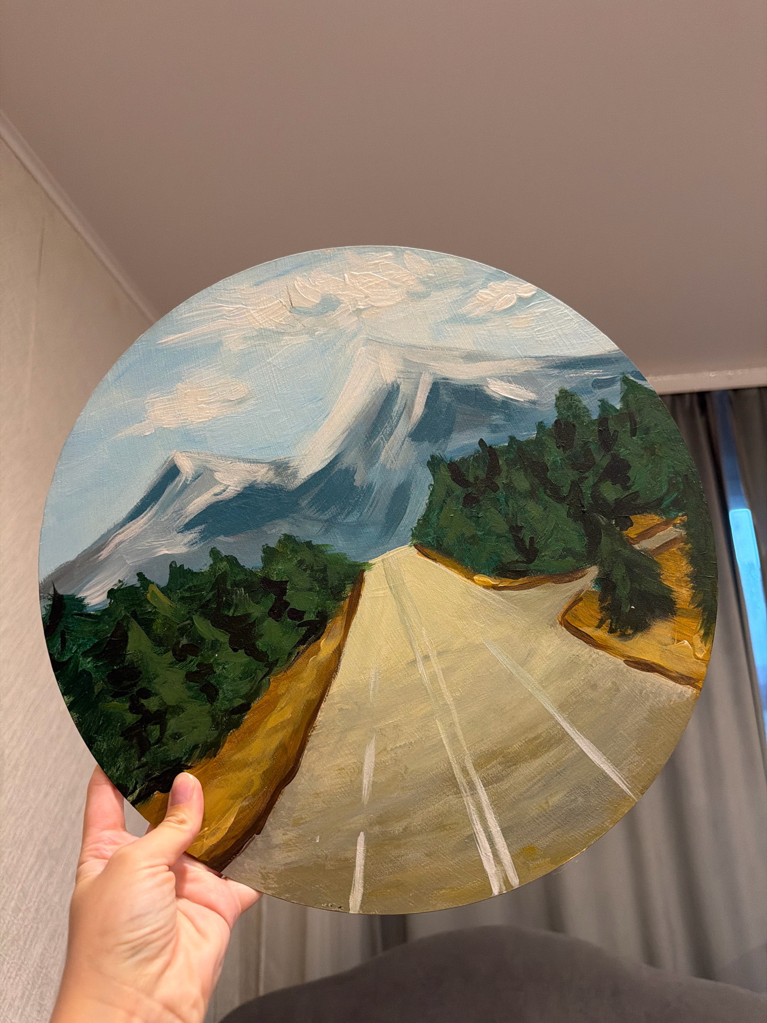 A circular painting on canvas depicts a mountain landscape with a road. The sky is a light blue with white clouds. In the background, snow-capped mountains are rendered in shades of white, grey, and blue. In the middle ground, a dense forest of dark green trees lines the sides of the road. The road itself is painted in yellow and white, with white lines indicating lanes. The foreground shows a hand holding the painting. The painting is indoors, with a wall visible in the background.