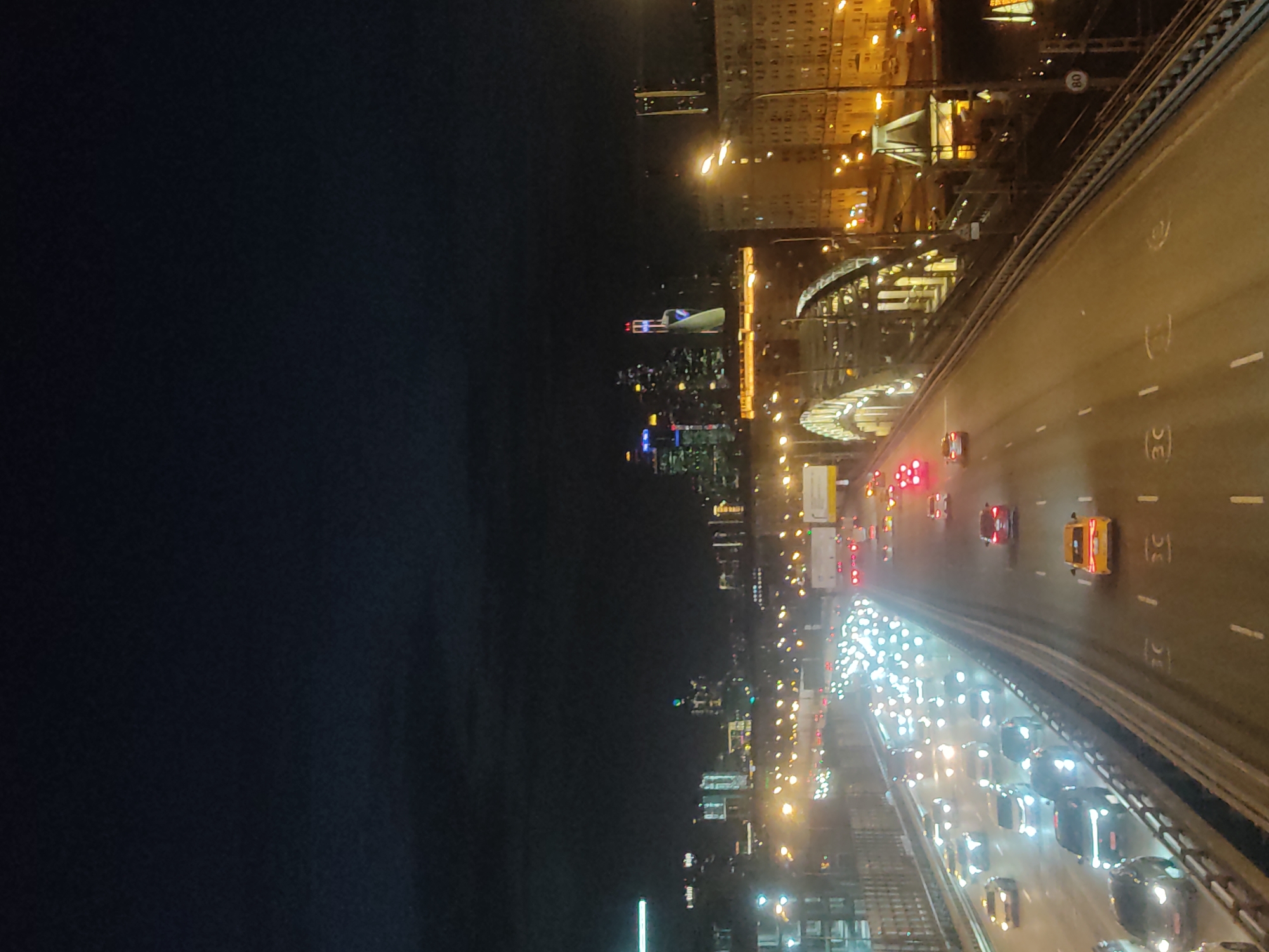 The image shows a nighttime view of Moscow, Russia, from a high vantage point, likely a bridge or tall building. The right side of the image features a multi-lane highway with traffic moving along it. Cars are visible, many with headlights on, suggesting a heavy flow of vehicles. Overhead street lighting illuminates the highway and the surrounding structures.
On the left, a skyline of Moscow can be seen, with identifiable buildings and landmarks lit up against the dark sky. Several skyscrapers stand out. A river appears to separate the highway from the city center. The area surrounding the highway and river is a mix of residential and commercial buildings.
