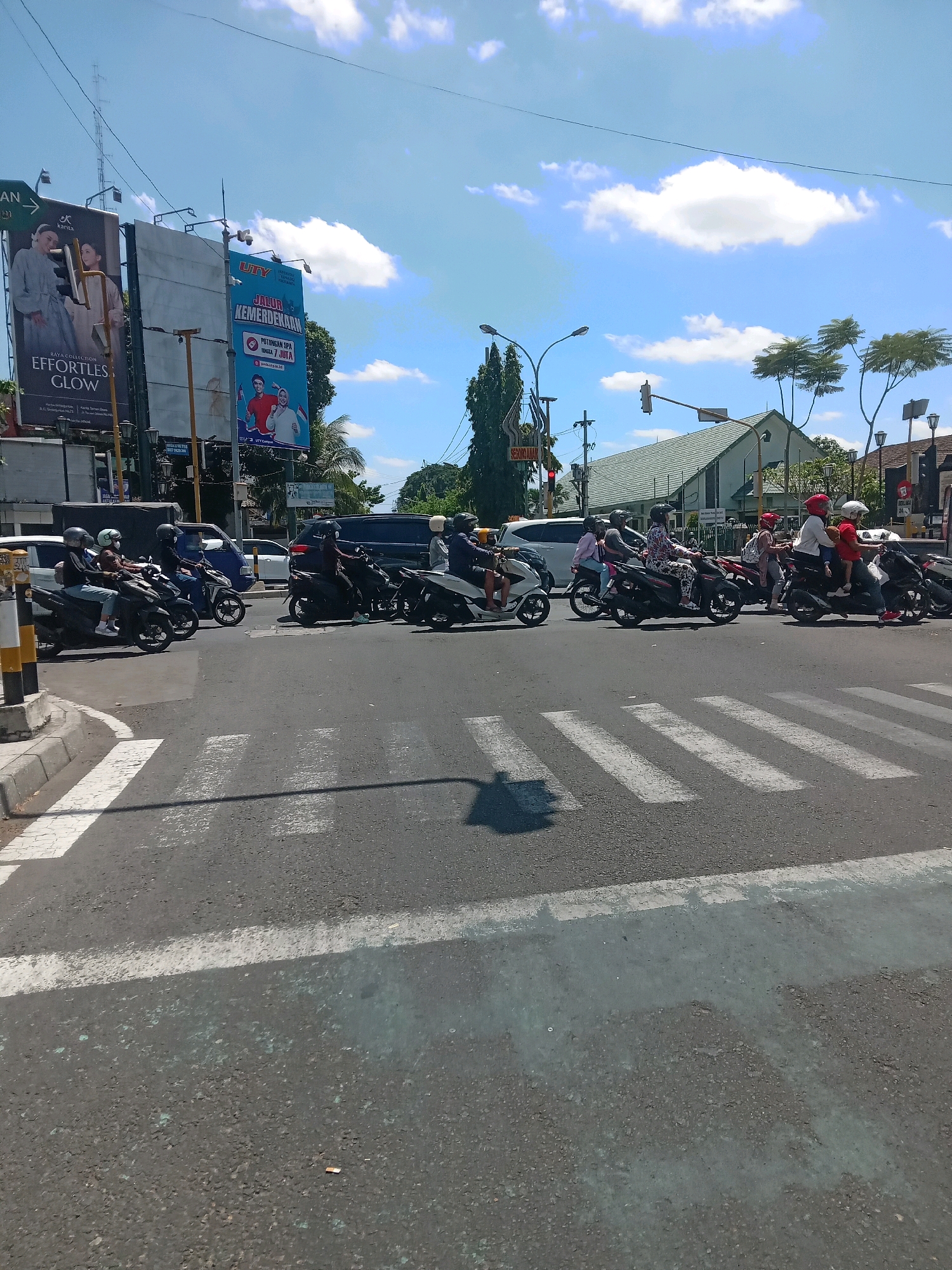 In Yogyakarta, Indonesia, on a bright, sunny day with scattered clouds, a traffic intersection is depicted. A busy flow of motorbikes is navigating the intersection, with riders wearing helmets. Some motorbikes are stopped at a pedestrian crossing, waiting for the light to change, while others are in motion. In the background, billboards with advertisements are visible, along with buildings and trees. The scene captures a typical urban streetscape in Yogyakarta, showcasing the prevalent use of motorbikes for transportation and the presence of commercial advertising.
