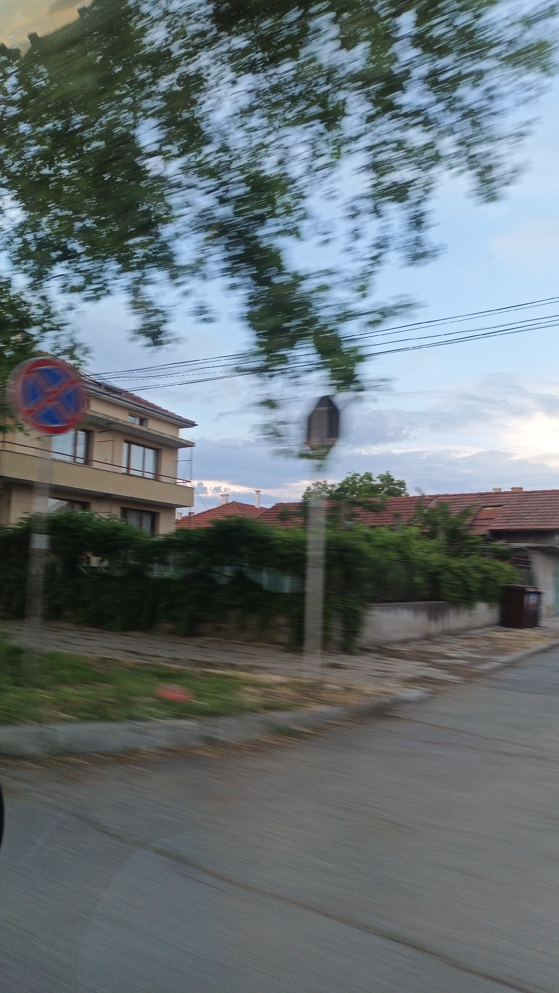 The image, likely taken from a moving vehicle, shows a residential street in Burgas, Bulgaria. The buildings include a two-story structure with a light-colored facade and reddish-brown roofs on other nearby houses. Lush green vegetation and trees are present, and a "No Stopping" sign is visible. The road is paved and appears relatively clean. The sky is partially cloudy. The blurriness of the image suggests motion. A dark-colored trash container can be seen on the side of the road. Electrical lines run across the sky.