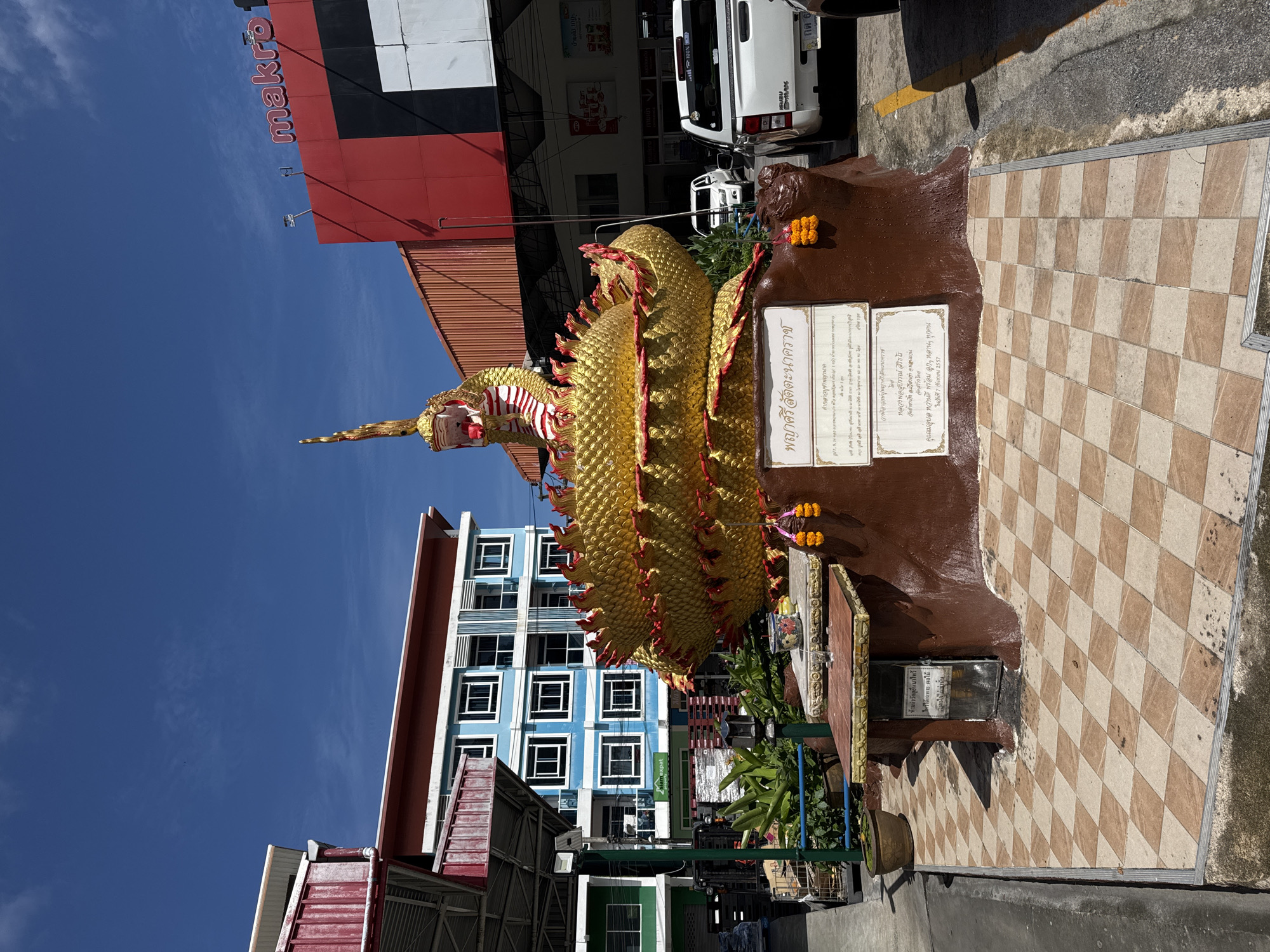 In Patong, Thailand, a large, golden serpent statue with red accents sits coiled in front of a brown, tree-like pedestal adorned with inscriptions and offerings. The statue is a prominent feature in the outdoor environment, with buildings and vehicles visible in the background. To the right, a white pickup truck is parked, and behind it, a Makro store is identifiable by its red and black signage. To the left, a blue apartment building with white-framed windows stands tall, with a red-roofed structure partially obscuring it. The ground is paved with a checkered pattern of beige and light brown tiles. The sky above is a clear, vibrant blue with a few wispy clouds.