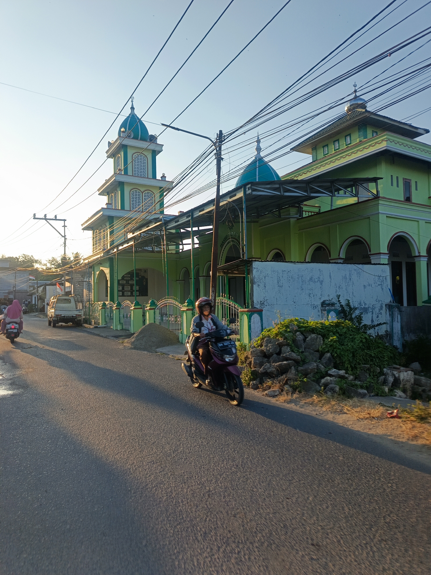 The image captures a street scene in Gorontalo, Indonesia, with a mosque prominently featured. The mosque is a light green color with a tall, square minaret topped by a blue dome. The main building also has a domed roof and several arched windows. A complex network of electrical wires crisscrosses the sky above the mosque.
In the foreground, a person is riding a dark purple scooter on an asphalt road. The rider is wearing a helmet and a light-colored jacket. Further down the road, another person on a scooter, wearing a pink hijab, is visible. A white pickup truck is parked on the side of the road. The sun is low in the sky, casting long shadows across the road and illuminating the scene with a warm, golden light. The environment appears to be a mix of residential and commercial buildings, with some vegetation visible on the side of the road.
