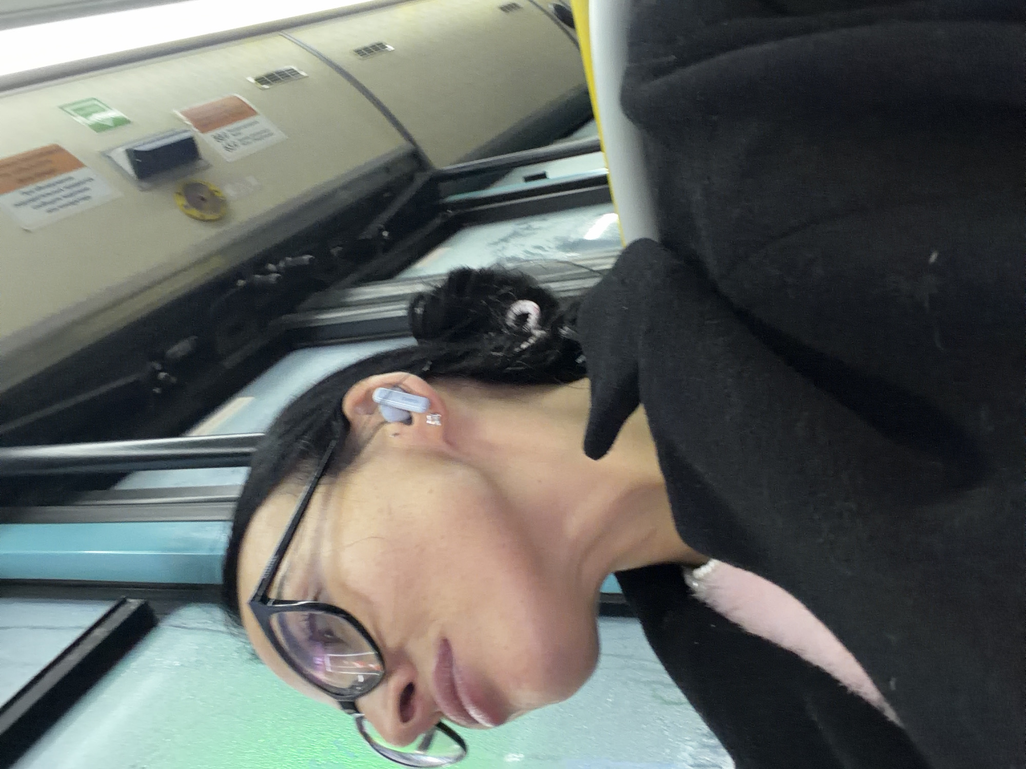 The image, rotated 90 degrees counter-clockwise, depicts a person's profile within what appears to be the interior of a public transport vehicle, likely a subway or train in Saint Petersburg, Russia.
The primary subject is a person whose right side of the face, neck, and shoulder are visible. They have dark hair pulled back into a bun or ponytail, secured with a small, white, possibly spiral-shaped hair tie. They wear black-framed glasses and a light grey earbud in their right ear, along with a small, square, shiny stud earring. The person is dressed in a black garment, possibly a jacket or hoodie, with a hint of light pink fabric visible beneath. Their expression appears neutral.
The setting is an indoor space, characterized by light-colored ceiling panels with air vents and various informational or warning signs. A distinctive yellow circular object, possibly an emergency button or control, is mounted on the panel. A narrow, light blue horizontal strip runs across the background, and reflections are visible on a lower surface. A yellow vertical pole is partially seen on the right.
The visible text on the signs is in Russian. One orange-brown label clearly shows "ПРЕДУПРЕЖДЕНИЕ!" (WARNING!) and another partially reads "ОБОРУДОВАН..." (EQUIPPED...). A green-colored icon is also present on one of the signs.
The person appears to be traveling, possibly listening to audio. The time of day and weather are not discernible from the interior shot. The unique angle suggests a candid shot, capturing a moment of everyday transit.