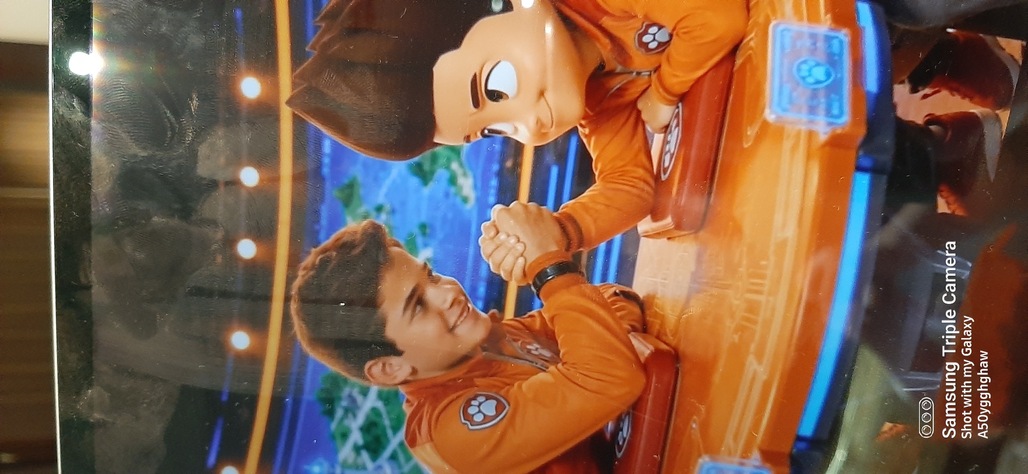 The image shows two young individuals arm wrestling on an orange table. One individual appears to be a real person with dark hair, wearing an orange jacket with a Paw Patrol logo. The other individual appears to be a character from Paw Patrol, possibly Ryder, with dark spiky hair and an orange jacket also featuring a Paw Patrol logo. Both are wearing red shoes. The background is a screen displaying a map with islands and a blue grid. The scene is lit by overhead lights. The lower portion of the image includes a watermark stating "Samsung Triple Camera Shot with my Galaxy A50yggghaw". There is no indication of this image being located in Dubai, United Arab Emirates.