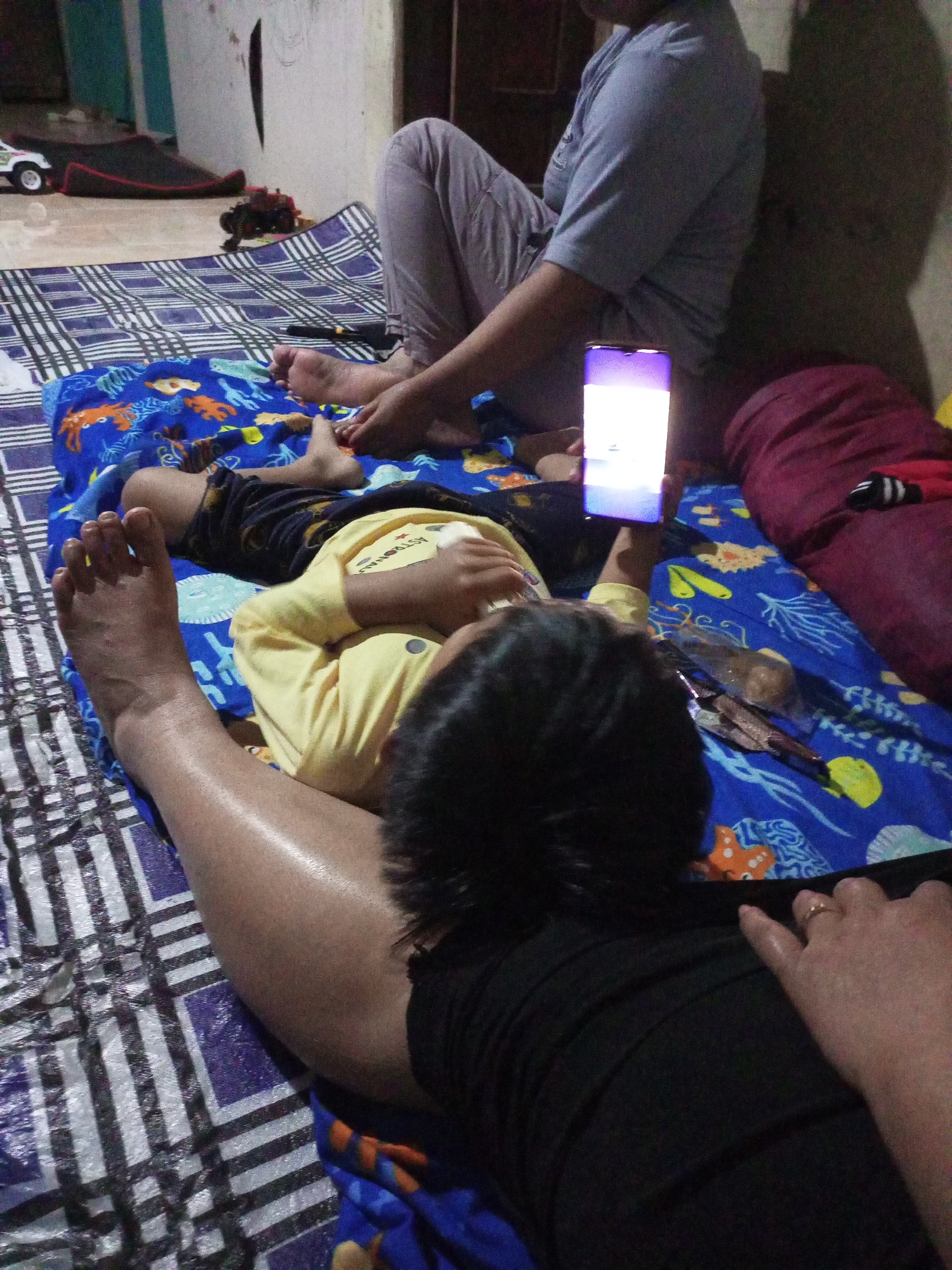 In a dimly lit room, possibly in Jatipurno, Indonesia, two individuals are visible. In the foreground, an adult's arm and hand, adorned with a ring, rest on a black garment. In the mid-ground, a child lies on their back, wearing a yellow shirt and dark shorts. The child is holding a smartphone, which is illuminated. Behind the child, another person, likely an adult, is seated and wearing light purple pants and a grey top. They are leaning against a wall. The individuals are on a patterned mat, with a blue blanket featuring marine life designs laid out underneath them. Various objects, including toys and what appears to be food items in plastic packaging, are scattered on the mat. The environment suggests a domestic setting.