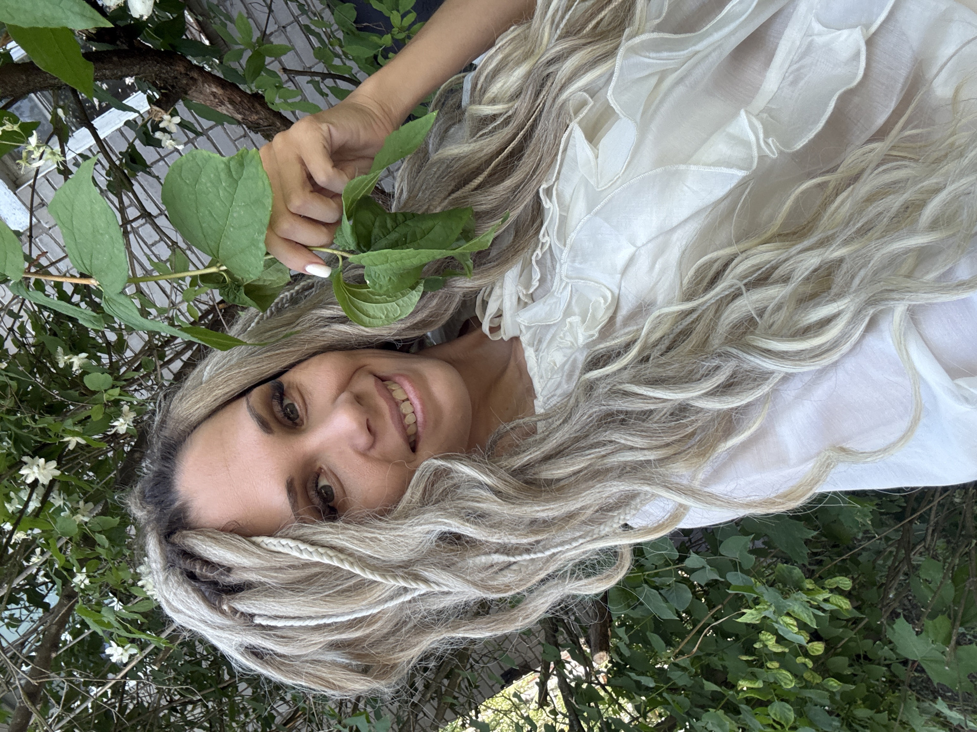 A woman with blonde dreadlocks is holding a green leaf and smiling at the camera. She is wearing a white ruffled shirt. The background is a blur of green foliage and a brick wall.