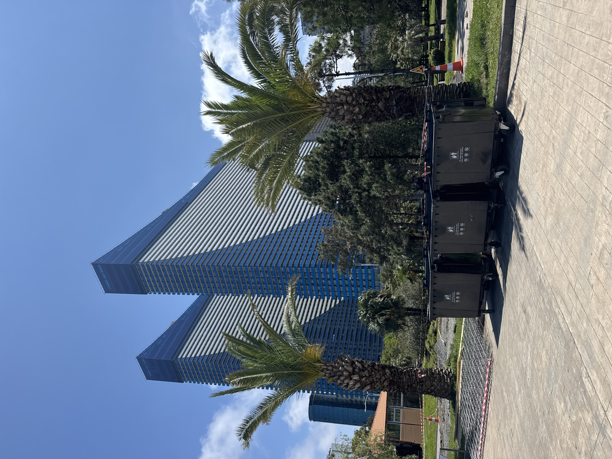 The image captures a scene in Batumi, Georgia, featuring modern architecture and urban elements. The primary structure is a tall, geometrically designed building with reflective glass facades, suggestive of a hotel or residential tower. Palm trees are situated in the foreground, partially obscuring the base of the building. To the right of the frame are several dark-colored refuse containers aligned along a paved walkway. A small area of landscaped greenery is present, with a safety cone visible near a temporary barrier. The sky is clear blue with scattered clouds. The overall impression is a sunny day in a developing urban area.