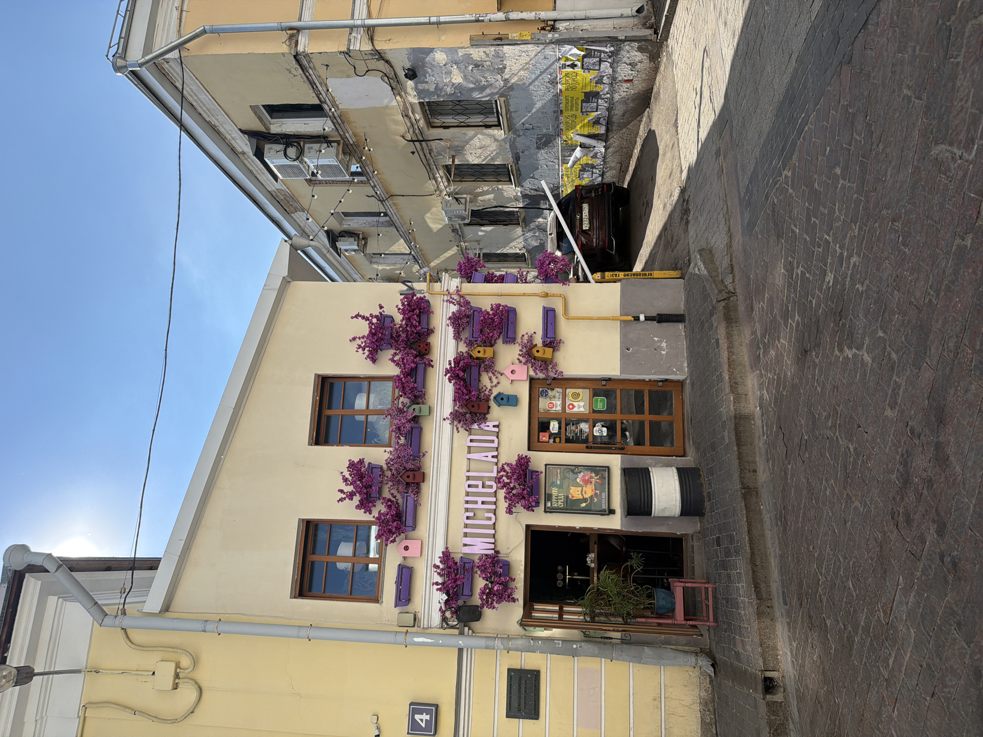 The image shows the facade of a building with the name "Michelada" displayed vertically on the front. The building is cream-colored with brown-framed windows. Purple flowers and colorful birdhouses adorn the facade around the windows and the name. To the right of the building, there is a narrow alleyway with a car parked at the end. The building next to Michelada has a weathered, gray exterior. The street appears to be paved with cobblestones. A black plaque and the number '4' are visible near the bottom left of the frame. The location is Moscow, Russia.