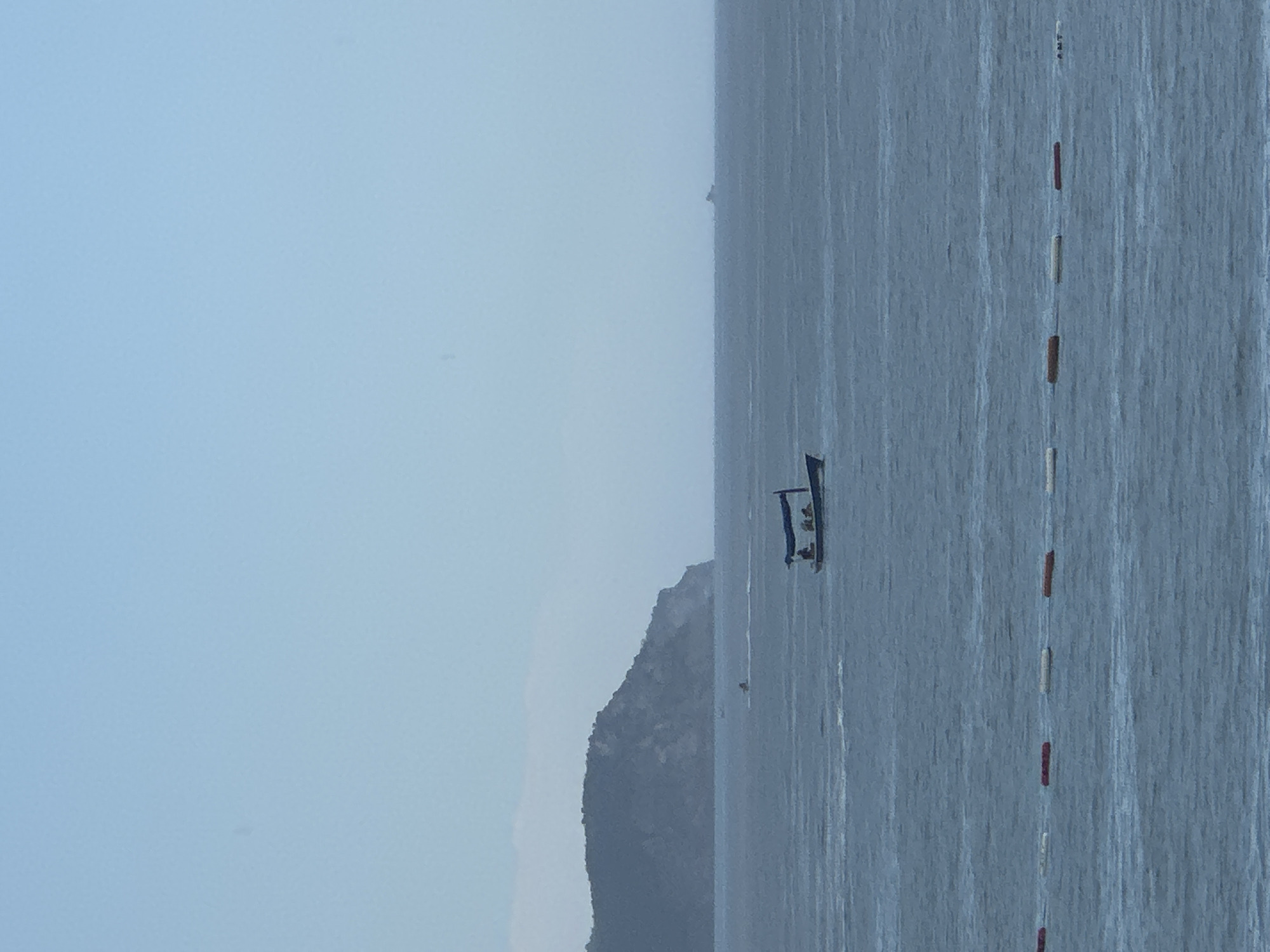 The image depicts a calm sea with a small boat in the distance. The boat appears to have a few people on board. A line of buoys marks a section of the water. In the background, a rocky, vegetated coastline rises from the sea, suggesting a coastal or island location. The sky is a pale blue, indicating it might be daytime, possibly early morning or late afternoon, with fair weather. The overall mood is peaceful and serene. There is no visible text in the image.