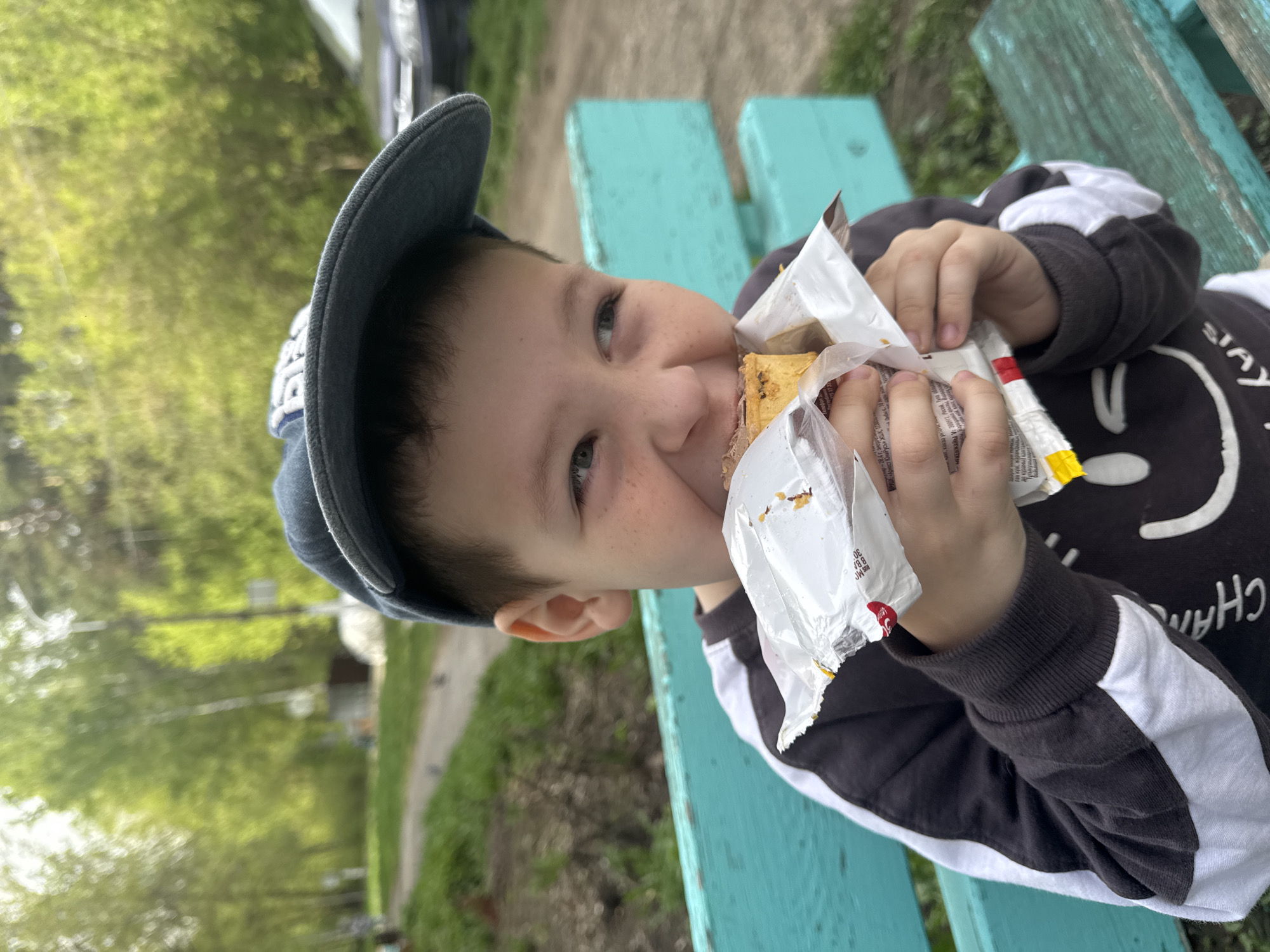 A young boy is sitting on a light blue bench outdoors, eating a sandwich wrapped in white packaging. He is wearing a dark gray baseball cap and a dark gray sweatshirt with white stripes and a white smiley face design. The packaging has some writing on it, including "Арнест", "ст ЮниРусь", "RUS", "BBA", and "30". The environment is green with trees in the background. The location is likely Chaikovsky, Russia, based on the Cyrillic text on the packaging.