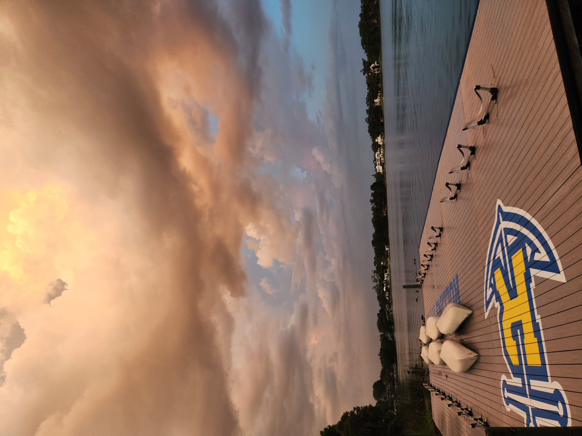 The image depicts a wooden dock on a lake, with a large university logo painted on it. The logo is blue and yellow, and appears to be for the University of Houston. In the background, there are trees and houses, suggesting a suburban or park-like setting. The sky is dramatic, with large, colorful clouds indicating either sunrise or sunset. Several white, boat-like objects are lined up on the dock. There are also several metal racks on the dock, possibly for storing boats. The scene is serene and tranquil, with the lake reflecting the sky. This scene is likely located at the University of Houston's boathouse on the West Campus.