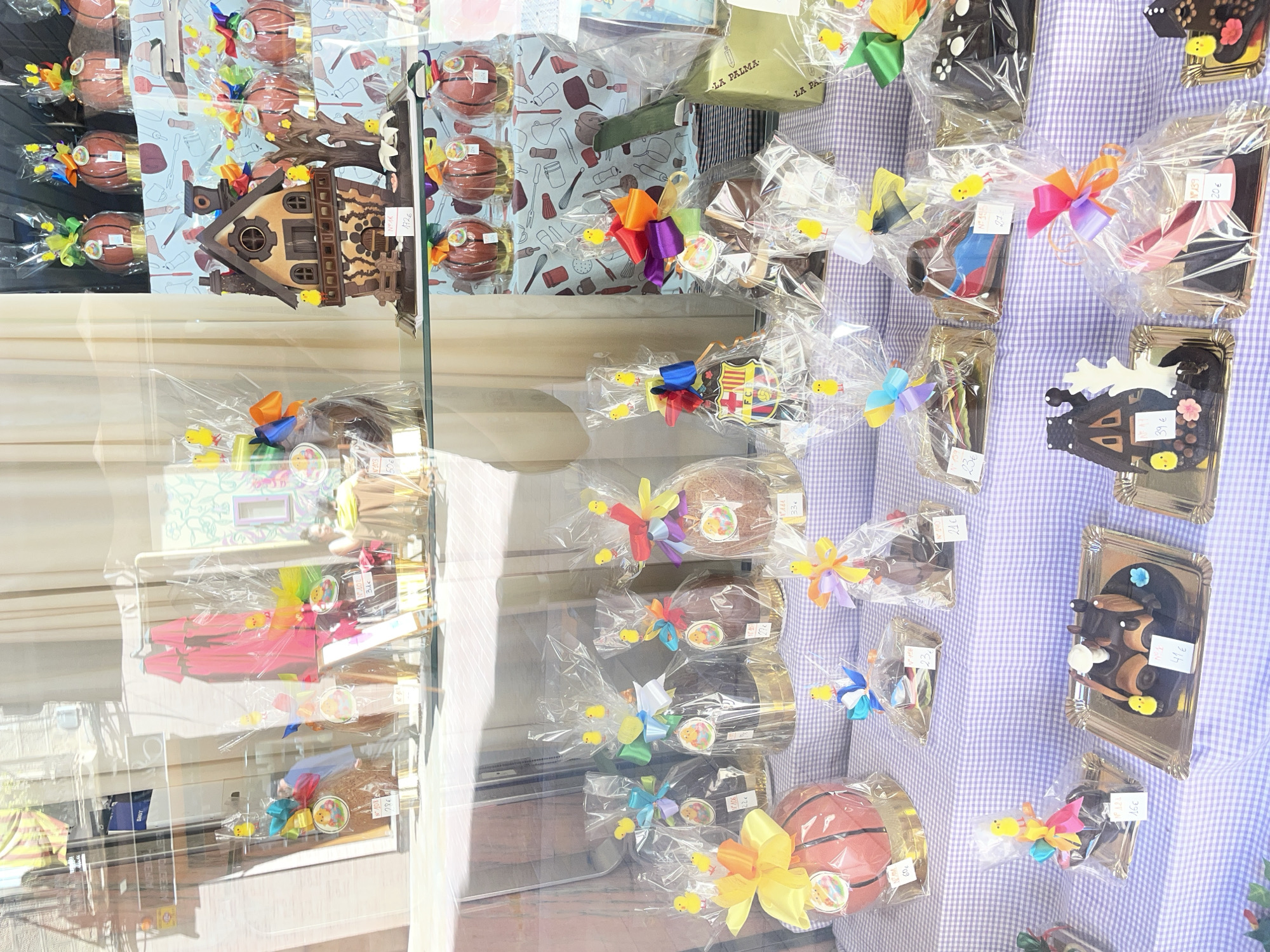 Here is a description of the image:
The image captures a window display of a pastry shop in Barcelona, Spain, likely preparing for the Easter season. The shop offers a variety of elaborately decorated chocolate sculptures and treats, indicative of the local custom to gift "Mona de Pascua" (Easter cake) to children.
The pastries are displayed against a background of a light blue and white checkered tablecloth, visible through a window that also reflects the outside urban scene, including building facades and sunlight.
The pastry items include:
* Chocolate Easter eggs, decorated and wrapped in transparent cellophane, some adorned with colorful ribbons and small chick decorations.
* A chocolate sculpture of a traditional Catalan country house, also known as "masia".
* Chocolate sculptures in the shape of basketballs, tied with large bows, showcasing the local passion for sports.
* Flat chocolate shapes that include depictions of traditional objects and designs, each placed on small golden trays and marked with a price tag.
The image presents a detailed view of the pastries themselves, their packaging, and pricing, as well as the surrounding environment. The scene emphasizes the cultural significance of these treats and the festive mood associated with Easter in Barcelona.
