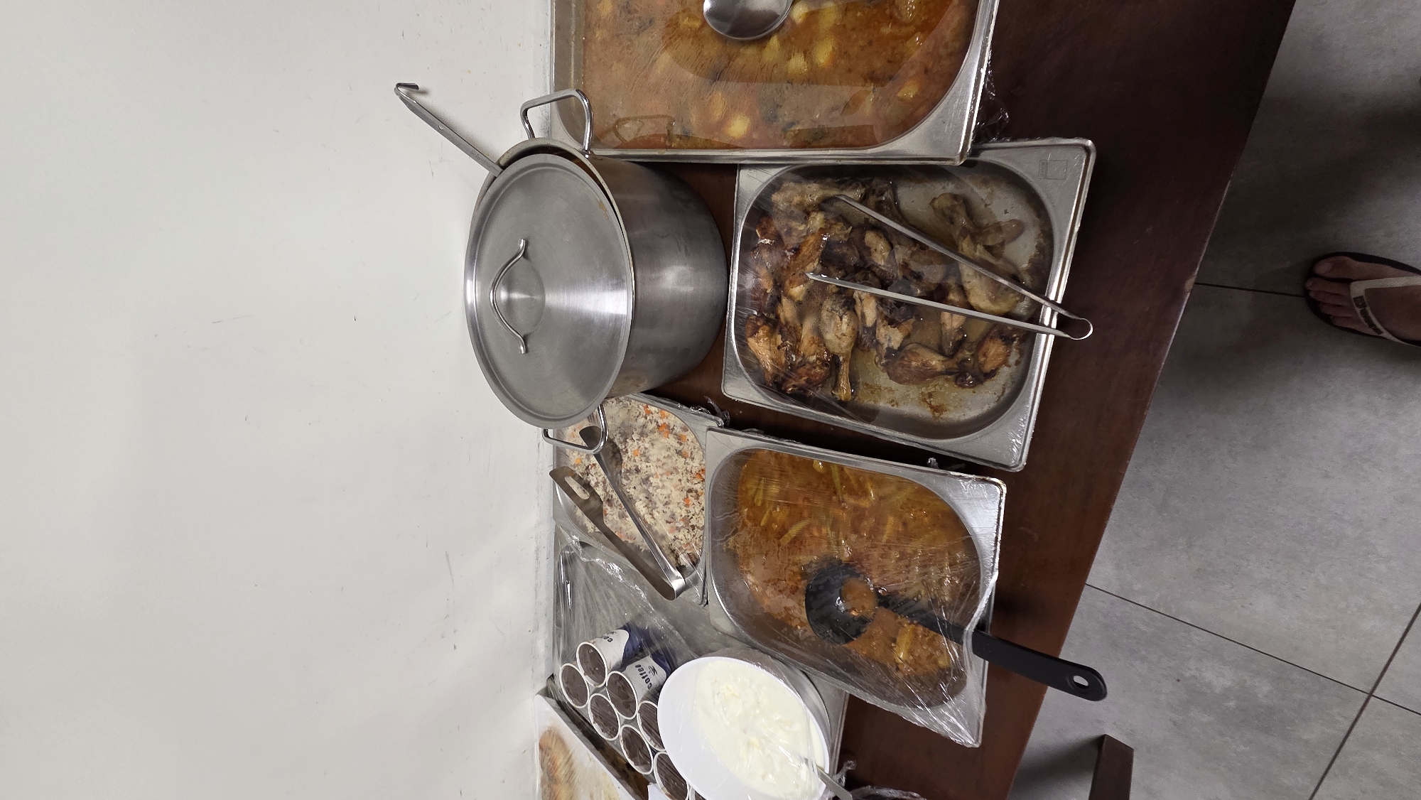 The image shows a buffet table in an indoor setting. The table is laden with various food items in silver serving trays and a large pot with a lid. The items include what appears to be a stew, roasted chicken, rice, and another stew-like dish with a large spoon. There are also small cups, a large bowl of what could be yogurt or a similar condiment, and a tray with a light-colored substance. Most of the trays are covered with plastic wrap.
A person's foot wearing a flip-flop is visible on the right side of the image, suggesting someone is standing nearby. The wall on the left is painted white. The table itself has a dark wood finish.
The specific location is not identifiable from the image, and the city of Unknown, Unknown, is not a real place.