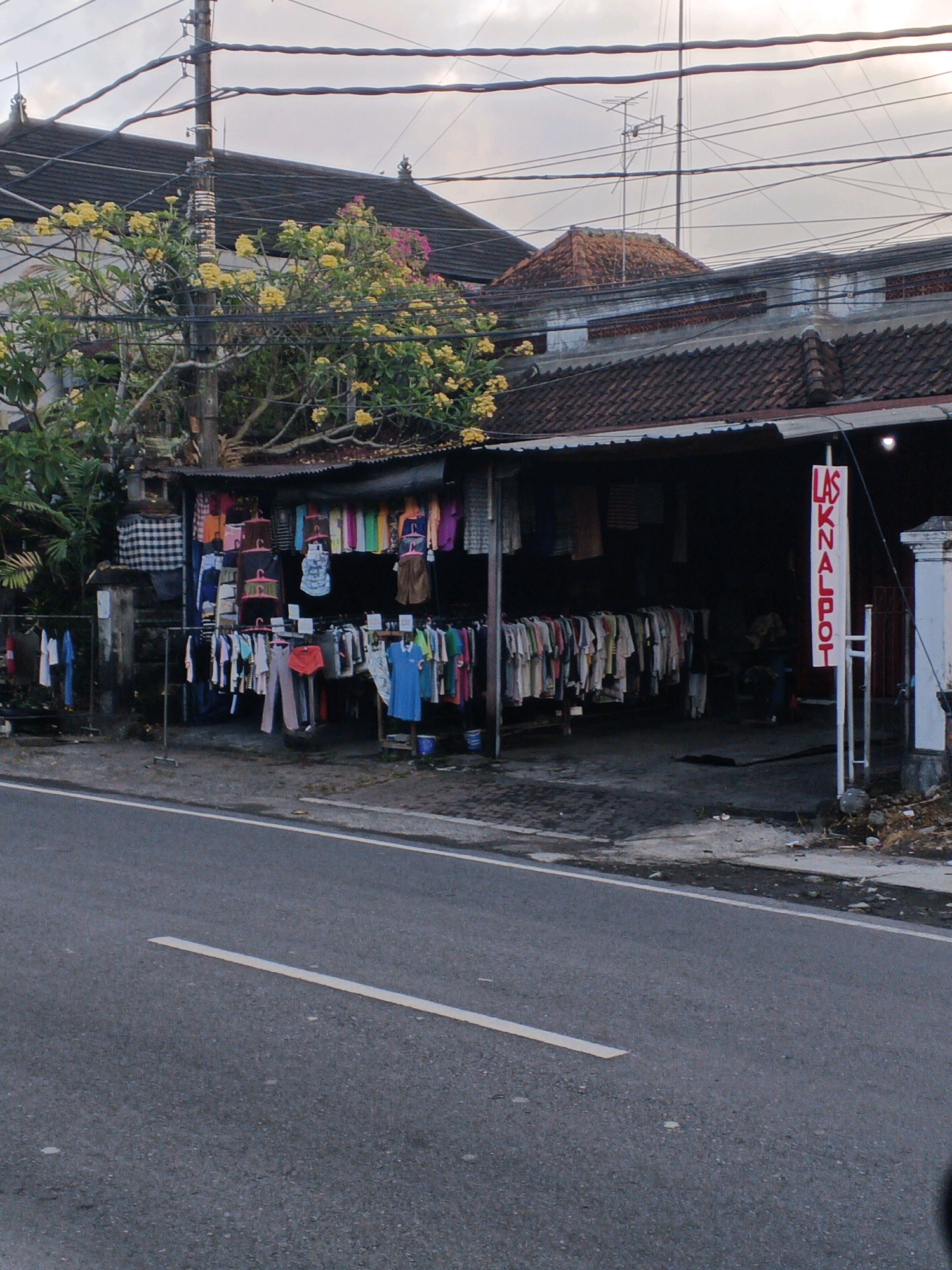 The image depicts a roadside clothing stall in Abiansemal, Indonesia. A variety of clothing items, including shirts, pants, and dresses, are displayed on racks and hangers. A red and white sign with text in a vertical orientation is visible on the right side of the stall. The stall is situated adjacent to a paved road with white lane markings. Behind the stall, a building with a tiled roof and an overhanging awning is visible, along with trees and power lines. The sky above is overcast. The scene appears to be captured during daylight hours.