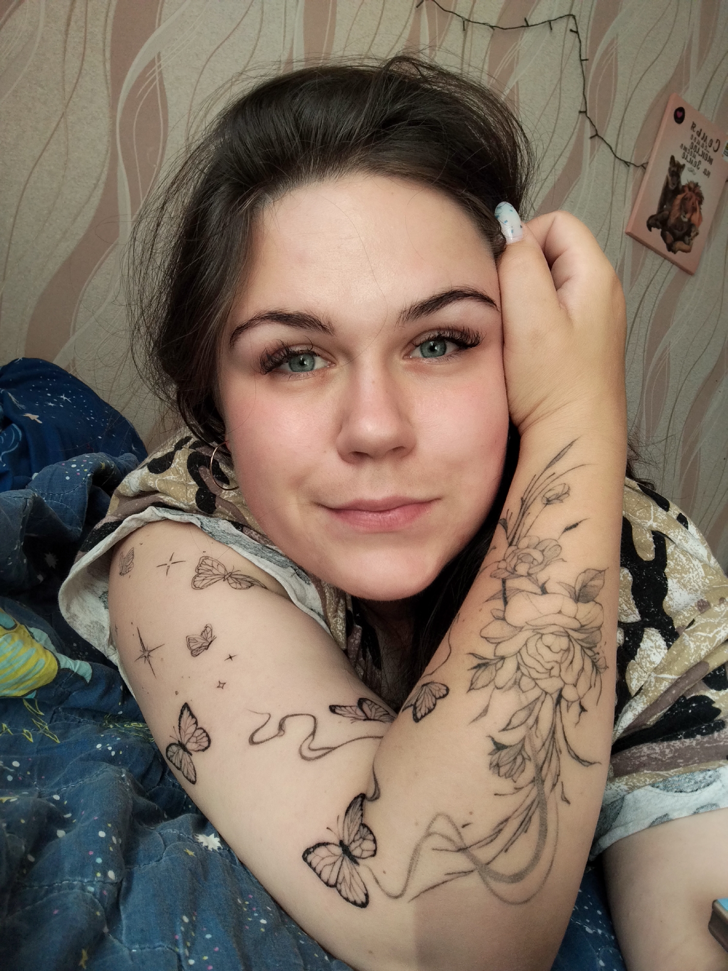 This media file is a close-up, eye-level selfie of a young woman with dark hair and blue eyes, lying down and resting her head on her hand. She is wearing a patterned top and appears to be indoors, possibly in a bedroom, given the presence of a blanket with a starry pattern and a wall with a textured wallpaper. The lighting suggests it is daytime.
The woman has several tattoos visible on her left arm. On her upper arm, there is a detailed floral tattoo, featuring roses and leaves. On her forearm and shoulder, there are smaller tattoos of butterflies and stars, as well as a swirling line. Her fingernails are decorated with a light blue and white pattern.
In the background, on the wall, there is a string of fairy lights and a small pink book with text in Cyrillic. The book appears to feature an illustration of two animals, possibly a wolf and a lion. The overall mood conveyed is relaxed and intimate. The woman has a slight smile on her face, and her gaze is directed towards the camera. The location context provided is Chaikovsky, Russia.