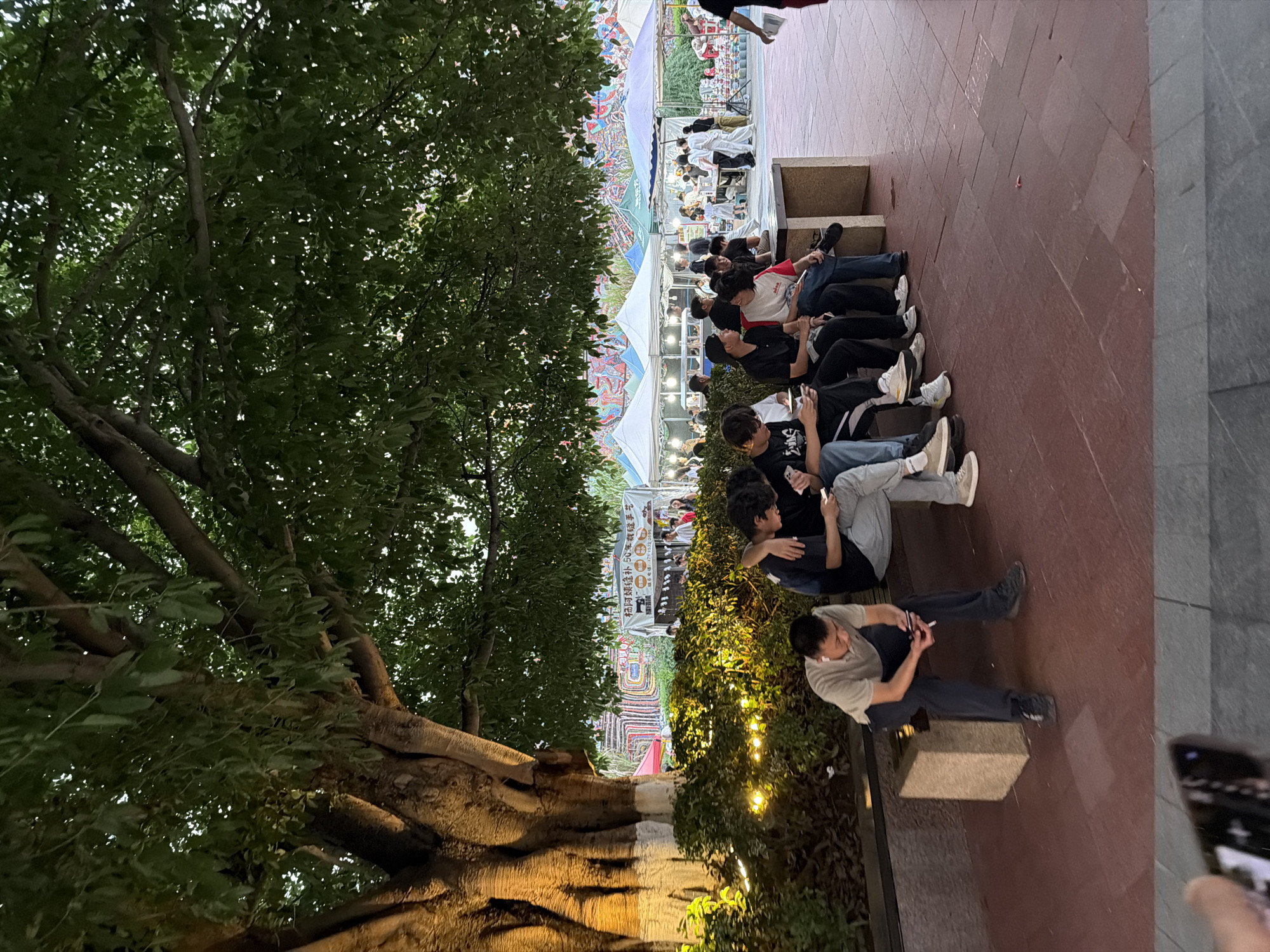 The image depicts an outdoor scene under a large, leafy tree. In the foreground, several young people are sitting on a concrete bench. Some are looking at their phones, while others are conversing. The bench is situated on a red tiled plaza. In the background, there are market stalls with tents, suggesting a street market or festival. The stalls display various goods, and some people are browsing. The lighting suggests it is late afternoon or early evening, with warm artificial lights illuminating the area around the bushes. The overall impression is a relaxed gathering in a public space. The location is specified as the Chongqing High-tech Zone, China.