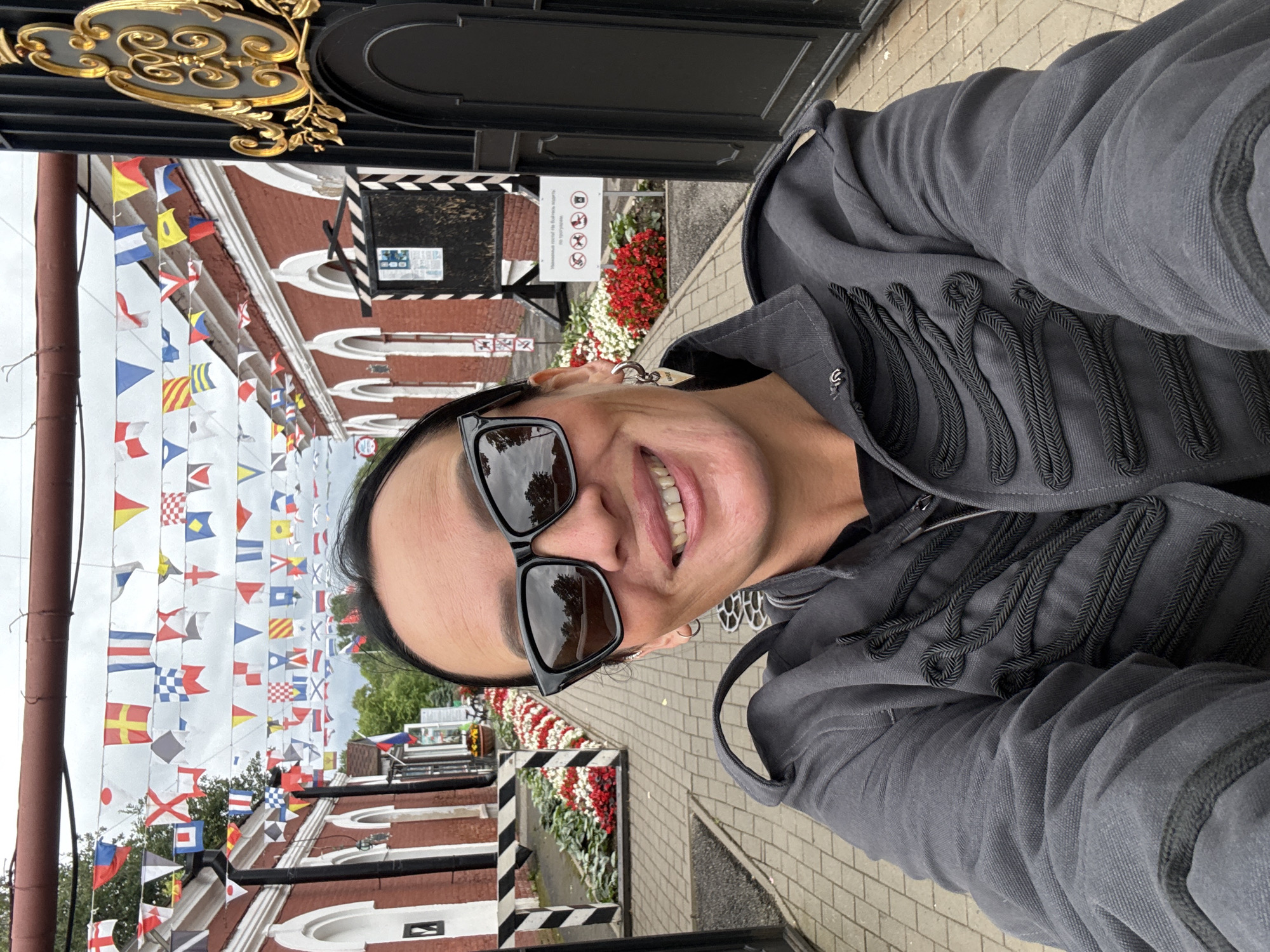 A woman with dark hair, sunglasses, and a gray jacket smiles at the camera while standing outside a building with red walls. She is wearing large, black, angular sunglasses and hoop earrings. The jacket is dark gray and has decorative black braiding on the front. In the background, a pathway is lined with flowerbeds, and a fence is visible. Above the pathway, numerous nautical flags are strung across wires. A large, ornate black and gold gate is to the right of the woman. The scene appears to be in Kronstadt, Russia, judging by the architecture and the style of the flags, which are typical of maritime settings.