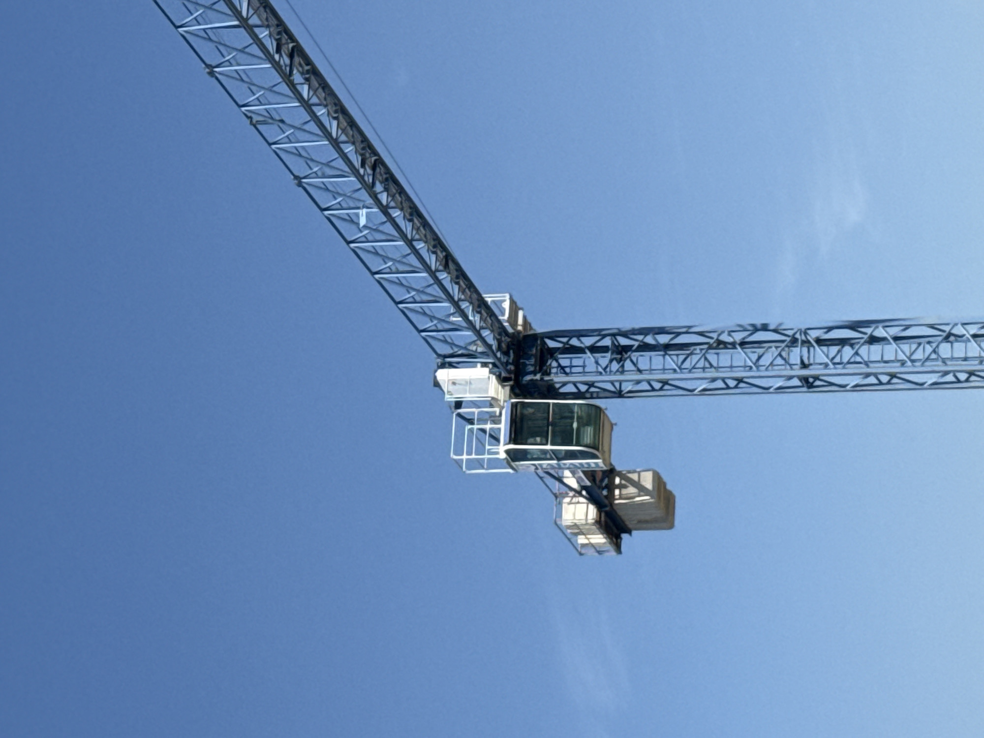 The image presents an upward view of a large, metallic construction crane set against a clear, bright blue sky. The primary subject is the intricate lattice structure of the crane, with its long boom extending diagonally from the upper left portion of the frame towards the center. From this central connection point, a shorter, horizontal jib extends towards the right.
A notable detail is the operator's control cabin, featuring prominent windows, positioned below the main jib and near the crane's pivot. Below and to the right of the cabin, additional structural elements or counterweights are visible. The crane's framework is composed of numerous interconnected metal trusses, characteristic of heavy construction equipment.
The sky is largely cloudless, save for a very faint, wispy cloud visible in the upper right, indicating fair weather and bright daylight conditions, likely during the mid-morning or afternoon. There is no specific activity depicted, suggesting the crane is stationary or paused in its operation. While the image itself contains no explicit location cues, this industrial scene is contextually set in Varna, Bulgaria. Close inspection of the operator's cabin reveals some barely discernible text or a logo on its side. No people are visible, and therefore no emotions are directly conveyed.