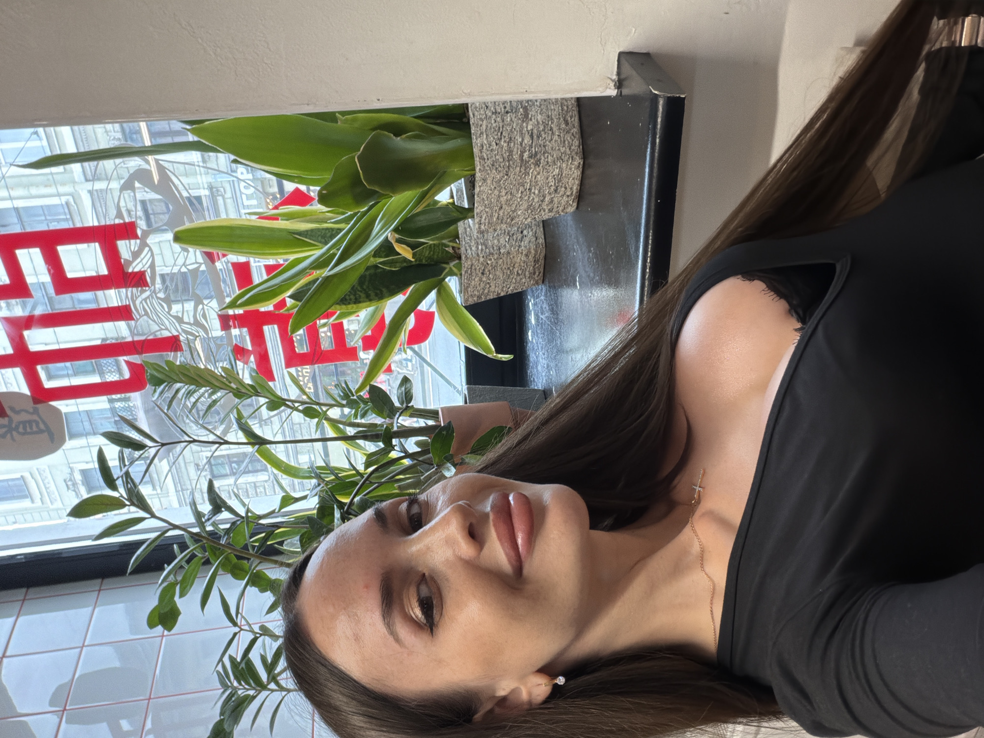 A young woman with long brown hair is taking a selfie in front of a window overlooking a city street. The woman is wearing a black top and a necklace with a cross pendant. The window has a red Chinese character sticker on it. In the foreground, there are several potted plants. The scene appears to be in Saint Petersburg, Russia, given the architectural style of the buildings visible through the window and the presence of Cyrillic text on a sign.
