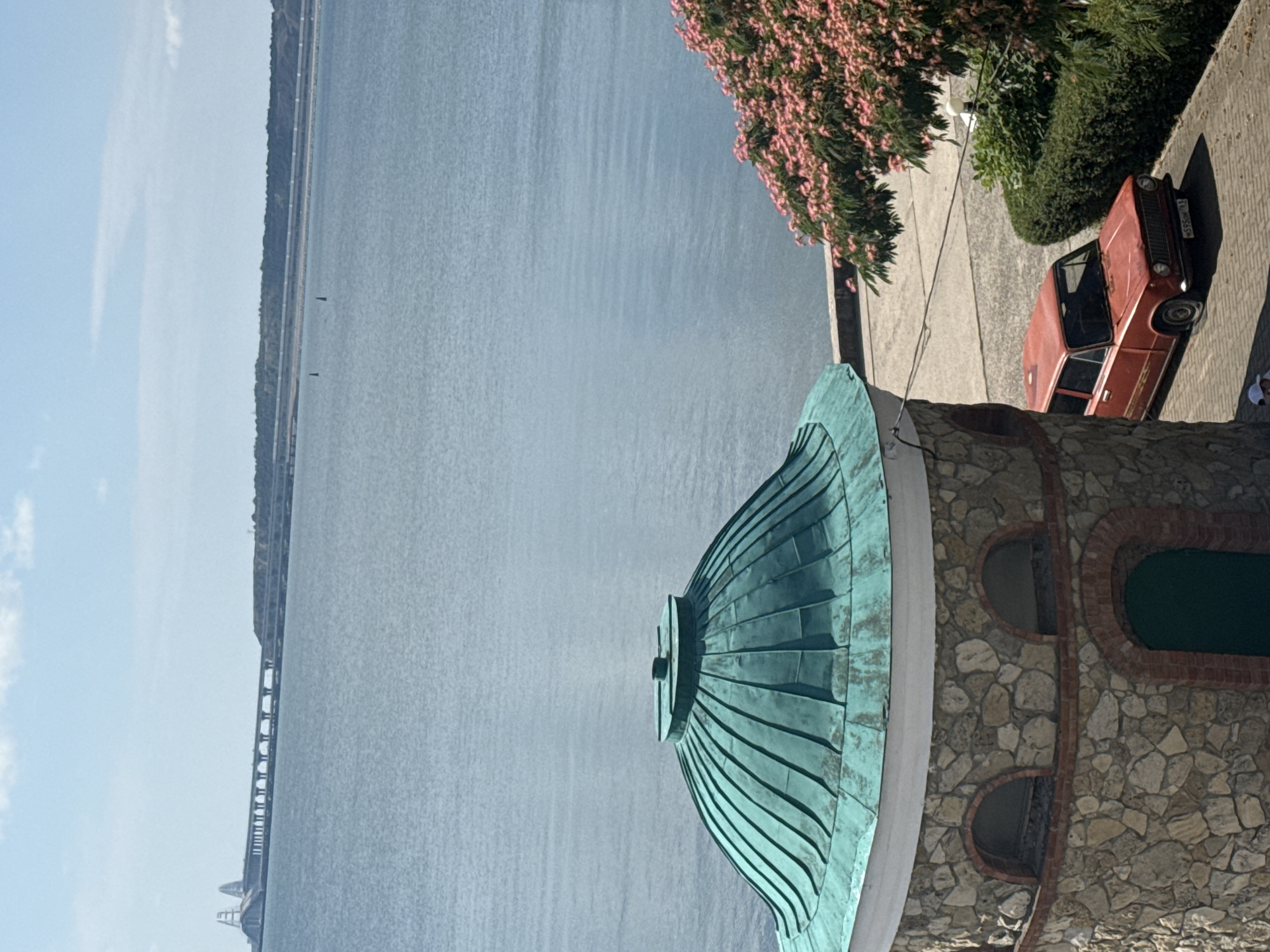 The image shows a view from a building with a stone facade and a green domed roof, overlooking a large body of water. In the distance, a long bridge spans the water, connecting land masses. On the right side of the image, a pink flowering tree and a red car are visible parked on a paved area. The car is a vintage model with a flat tire. The overall scene suggests a coastal location.