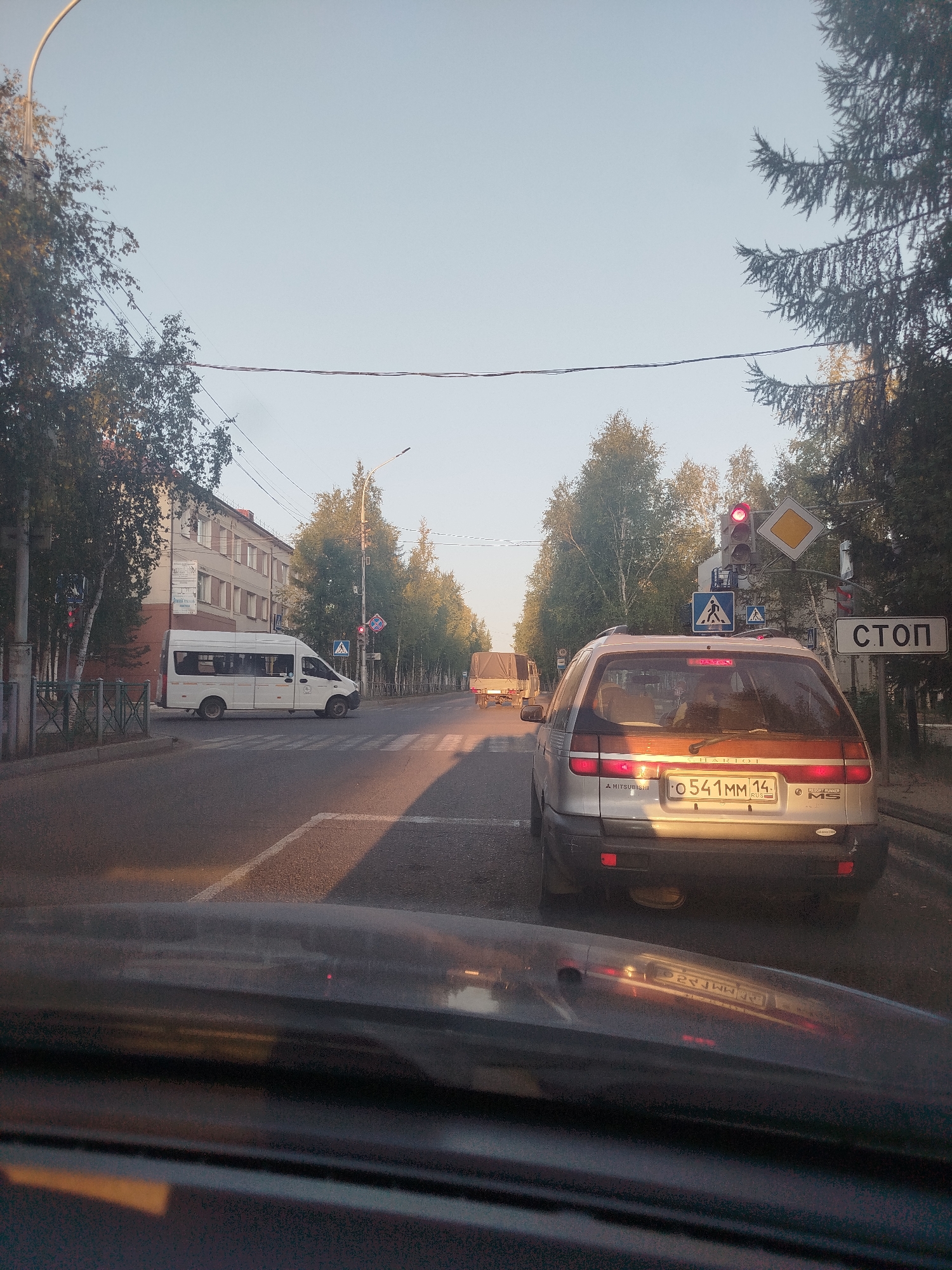 A view from inside a vehicle looking out at a city street in Mirny, Russia, during daylight hours. The street is paved, with a pedestrian crosswalk visible. On the left side of the street, a multi-story building with a light-colored facade and numerous windows stands behind a row of trees and a metal fence. To the right of the building, a white passenger van is parked. Further down the street, a silver station wagon is stopped at a traffic light. A truck with a canvas cover is also visible further in the distance. A traffic light with a red signal is illuminated, and various road signs are present, including a diamond-shaped yield sign, a pedestrian crossing sign, and a "STOP" sign. Trees line both sides of the street, and the sky is clear and light blue. The scene appears to be a typical urban streetscape during the day.