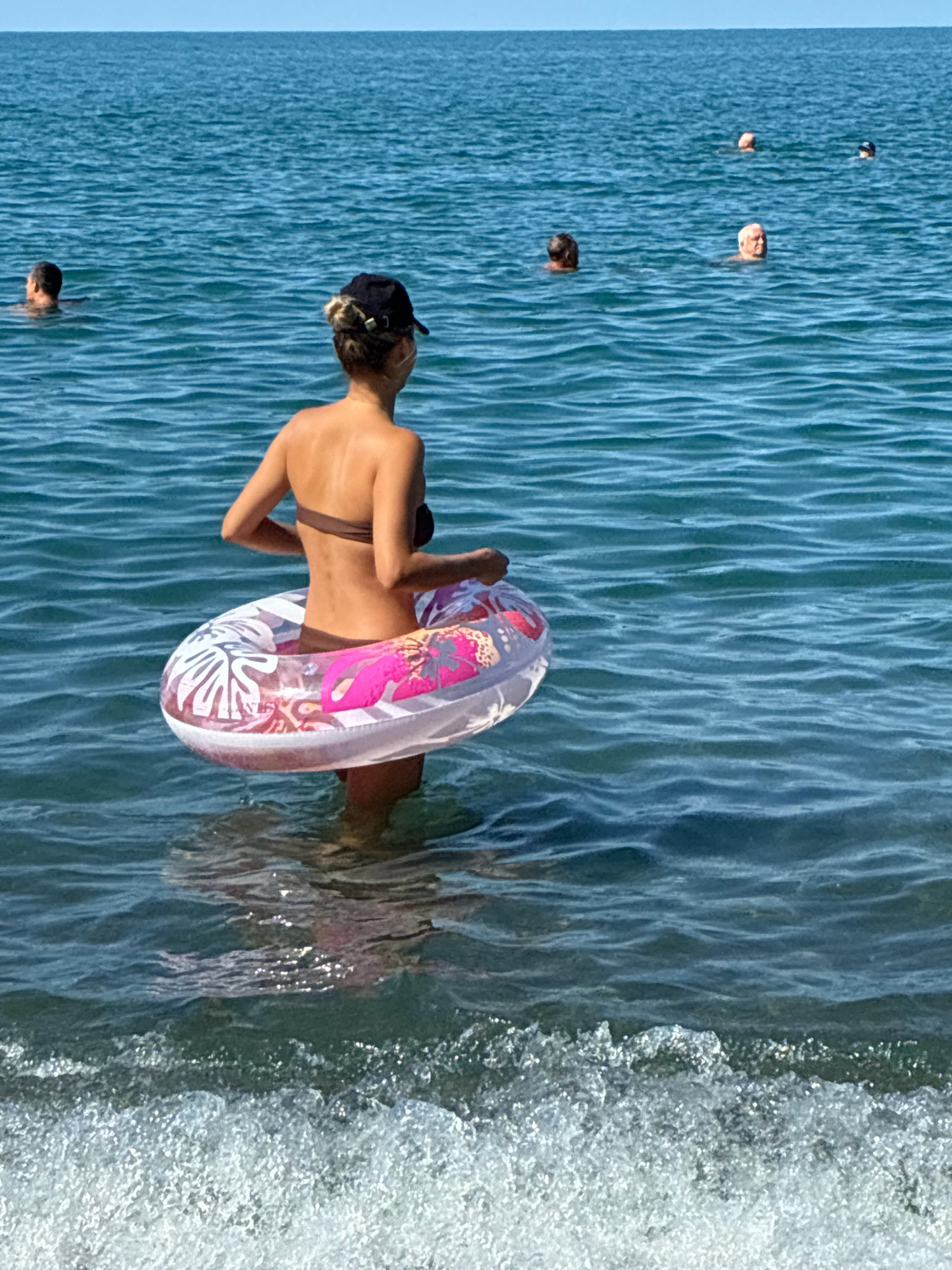 A woman wearing a brown bikini and a black baseball cap stands in the sea, holding a circular inflatable ring with a pink flamingo and leaf pattern. The ring is around her waist. The water is calm and blue. In the background, several people are swimming or wading in the sea. The scene is likely on a beach in Sochi, Russia, given the clear blue water and sunny sky.