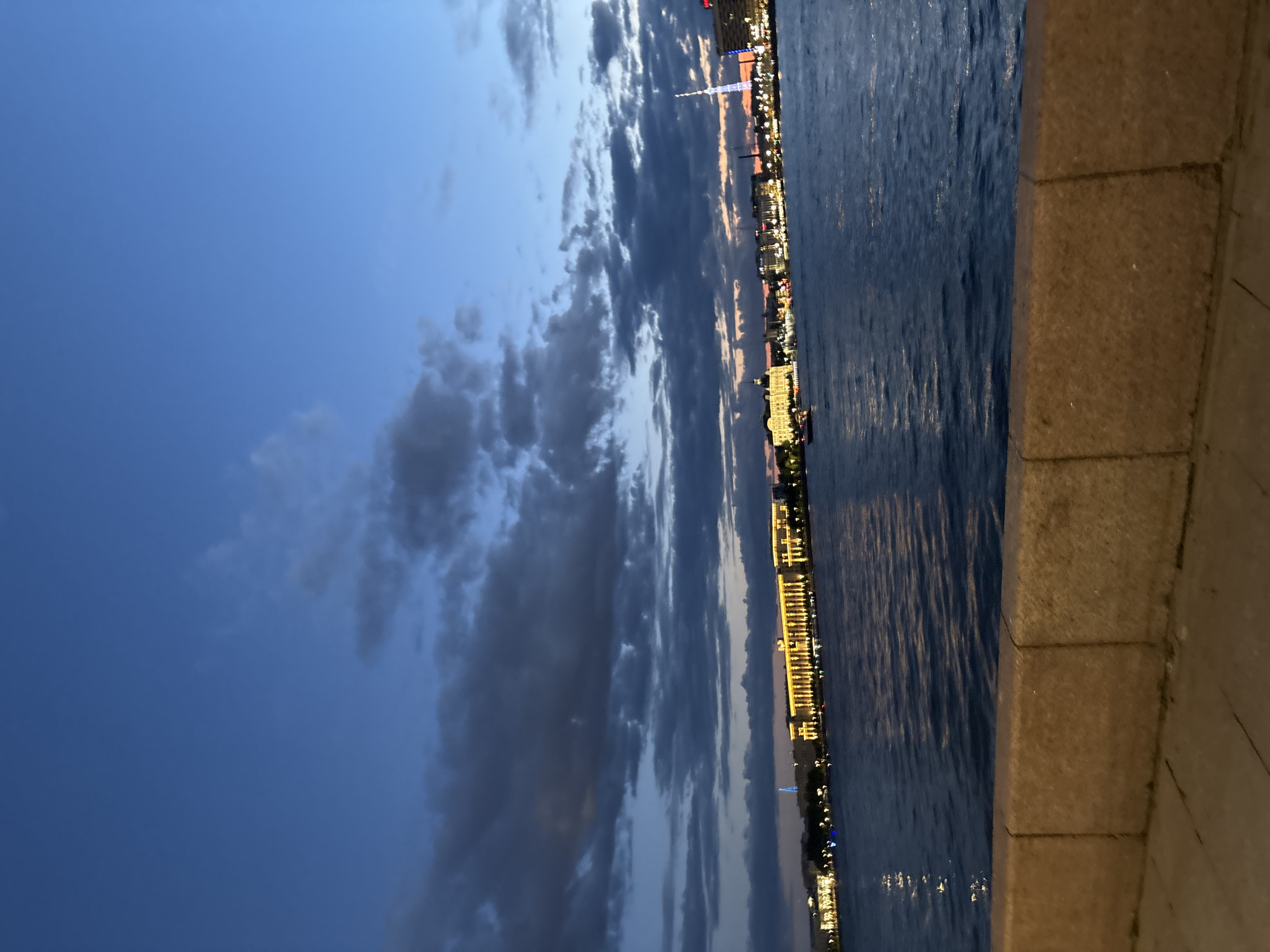 The image shows a twilight view of the Neva River in Saint Petersburg, Russia. On the far bank, a long, illuminated building with numerous windows is visible, likely a historical structure. Several boats are on the river, and lights from the city buildings reflect on the water. The sky is a deep blue with scattered clouds, indicating the transition from day to night. In the foreground, a stone embankment lines the river, with a paved walkway behind it. A tall, slender tower with lights, possibly a communication tower, is also visible in the distance. The scene is peaceful and serene, showcasing the beauty of Saint Petersburg at dusk.