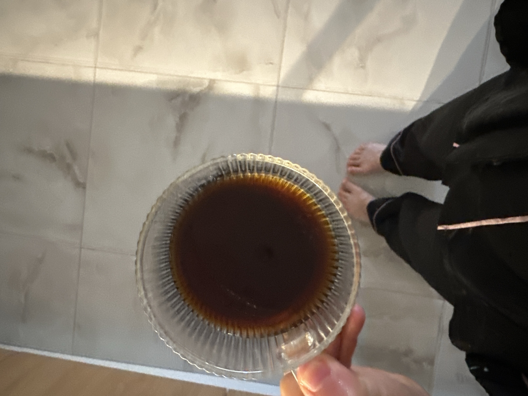 The image presents a top-down view of an indoor scene in Budva, Montenegro. The primary subject is a person, partially visible, holding a clear, ribbed glass cup filled with a dark beverage, likely coffee or tea. The person's bare feet are on a light-colored, marble-patterned tiled floor, suggesting a relaxed domestic or hotel setting. They are wearing dark, loose-fitting clothing, possibly pajamas or loungewear. A prominent shadow is cast across the tiled floor, indicating natural light, possibly sunlight, entering the room from an angle. The overall impression is of a calm, quiet moment, perhaps during a morning routine. No visible text is present in the image.