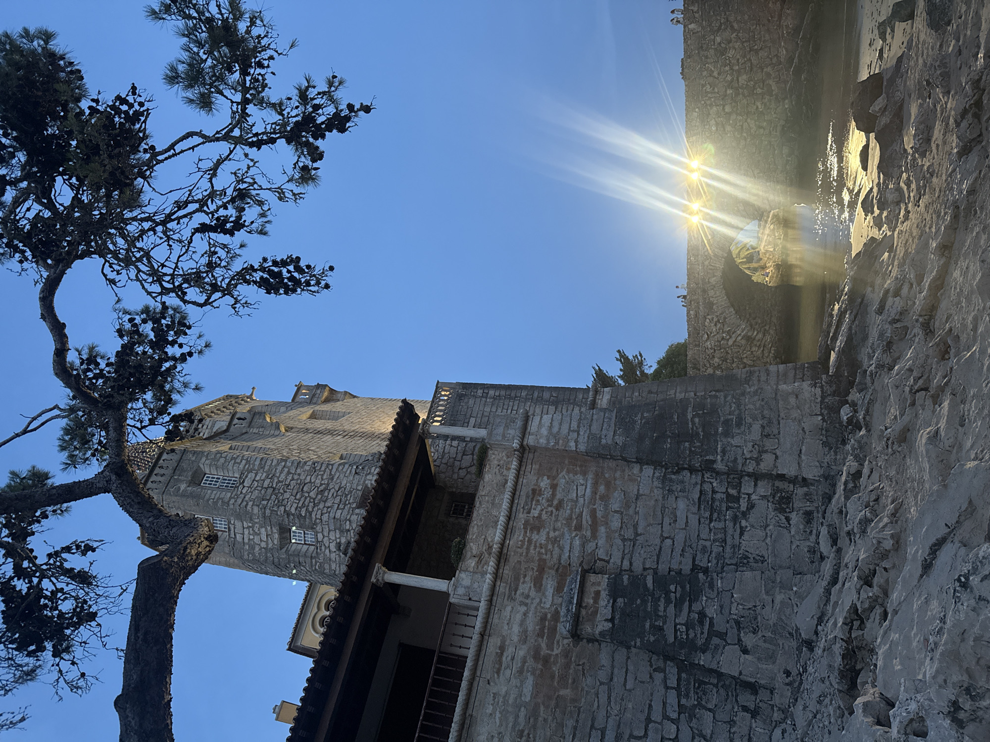 The image depicts a stone castle tower and adjacent wall in the foreground, with a stone arched bridge illuminated by bright lights in the midground. The castle structure is made of rough-hewn stone blocks, with a tiled roof and windows. A balcony with columns is visible on the left side of the castle. A pine tree branch hangs from the top left corner of the image. The bridge spans a body of water, and several small figures of people can be seen standing on and near the bridge. The water reflects the bright lights from the bridge, creating a shimmering effect. The sky is a clear blue, indicative of dusk or dawn. The location appears to be the Boca do Inferno in Cascais, Portugal, a natural sea arch known for its dramatic rock formations and crashing waves, which is often illuminated at night. The scene is captured from a rocky shoreline.