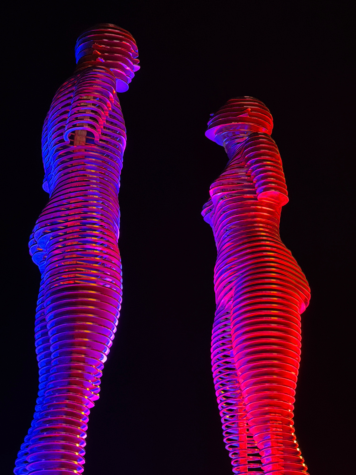 The image shows two illuminated, segmented humanoid sculptures against a dark background. The sculptures are constructed from horizontal, overlapping rings that create a layered, abstract human form. The sculpture on the left is predominantly lit with blue and purple light, while the sculpture on the right is primarily illuminated with red and pink light. The sculptures appear to be positioned facing each other. These are the Ali and Nino sculptures, located in Batumi, Georgia, on the Black Sea coast. The dynamic lighting suggests the sculptures are illuminated at night.