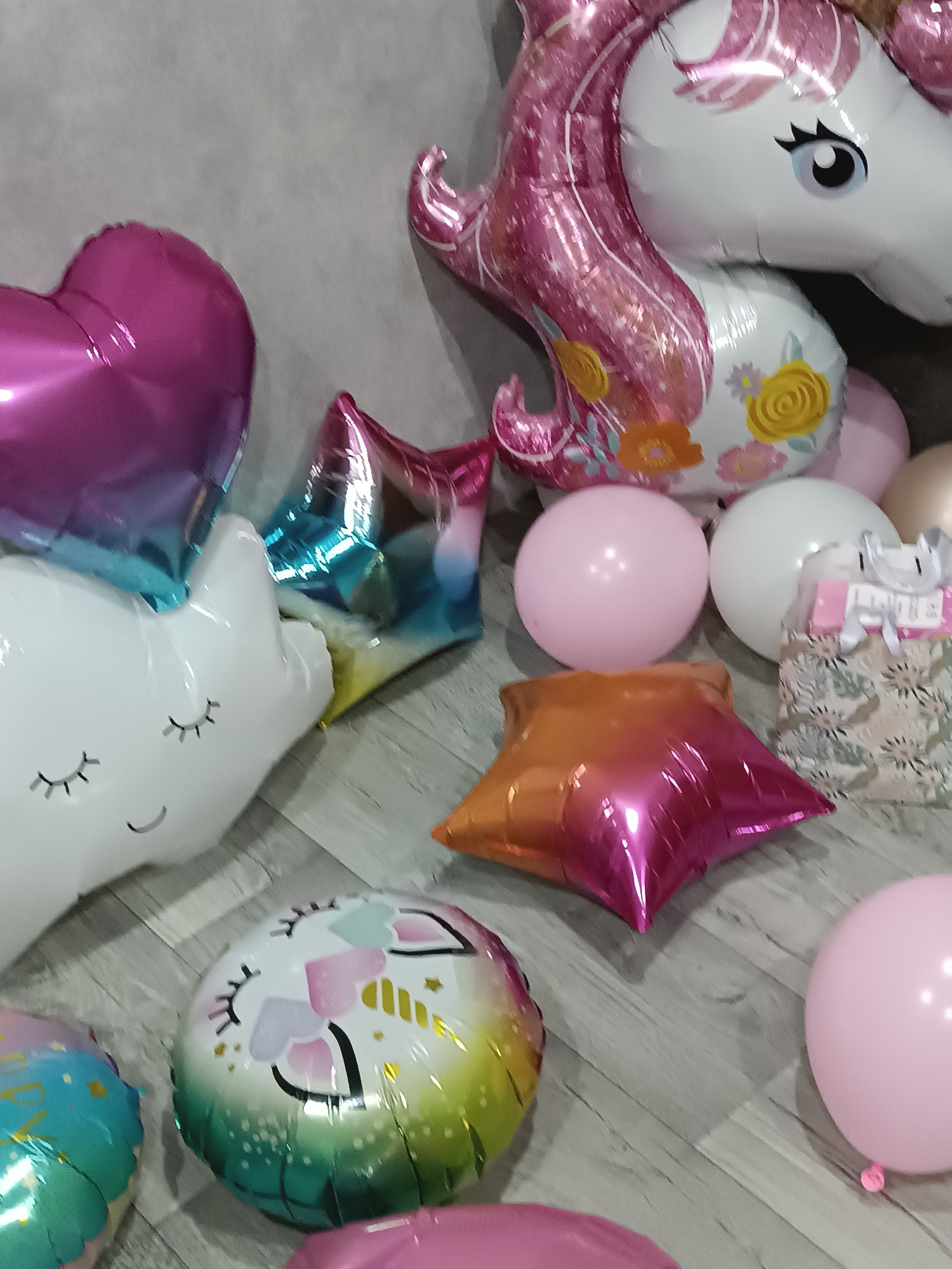 The scene depicts an indoor setting, likely a room, in Unknown, Unknown. A collection of assorted balloons and one gift bag are arranged on a light gray wooden floor, with a plain light gray wall visible in the background. Dominant objects include a large white foil unicorn balloon with a pink mane and floral accents, a large pink and blue gradient foil heart balloon, and a large white foil cloud balloon with a face. Additionally, there are two medium-sized foil star balloons, one with a blue-to-pink gradient and another with an orange-to-pink gradient. A round foil balloon featuring a unicorn face with a rainbow gradient mane is also present. Multiple pink and white spherical latex balloons, along with one gold/beige latex balloon, are interspersed among the foil balloons. A small patterned gift bag with a pink ribbon and a white decorative element is positioned near a white latex balloon. No individuals or active interactions are depicted.