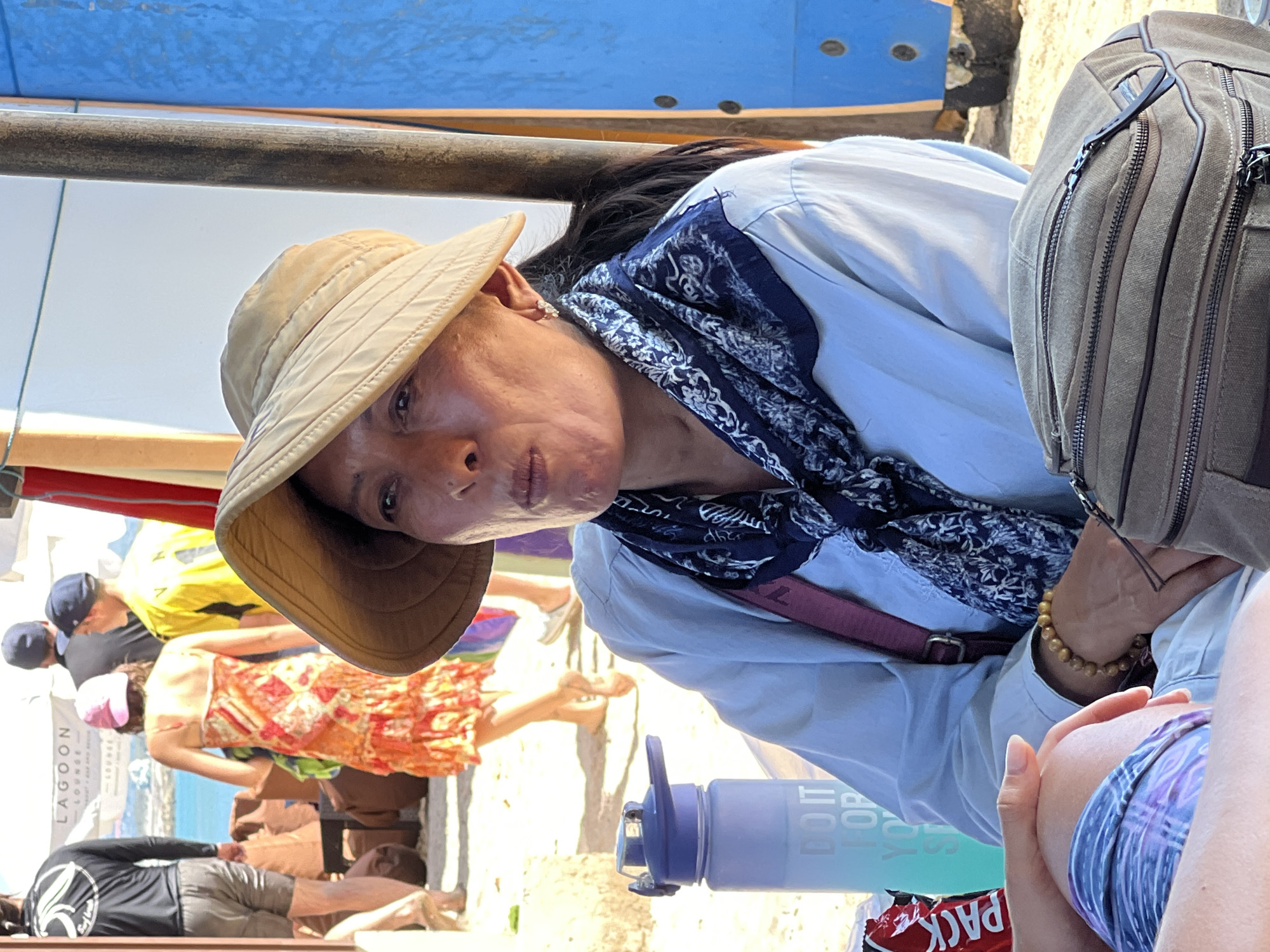 A medium shot captures a woman with dark hair, wearing a light blue button-down shirt, a dark blue patterned scarf, and a light tan wide-brimmed hat. She is looking slightly to her left with her mouth open. She wears a beaded bracelet on her left wrist and a thin strap across her chest. To her left is a blue and green water bottle with the words "DO IT FOR YO" visible. In the foreground, a portion of a grey canvas bag and a person's legs in purple and blue patterned shorts are visible. In the background, other people are seen in casual beach attire, and a sign that reads "LAGOON LOUNGE" is visible. The scene appears to be an outdoor setting, possibly a beach or a coastal establishment, consistent with a location in Kuta, Indonesia.