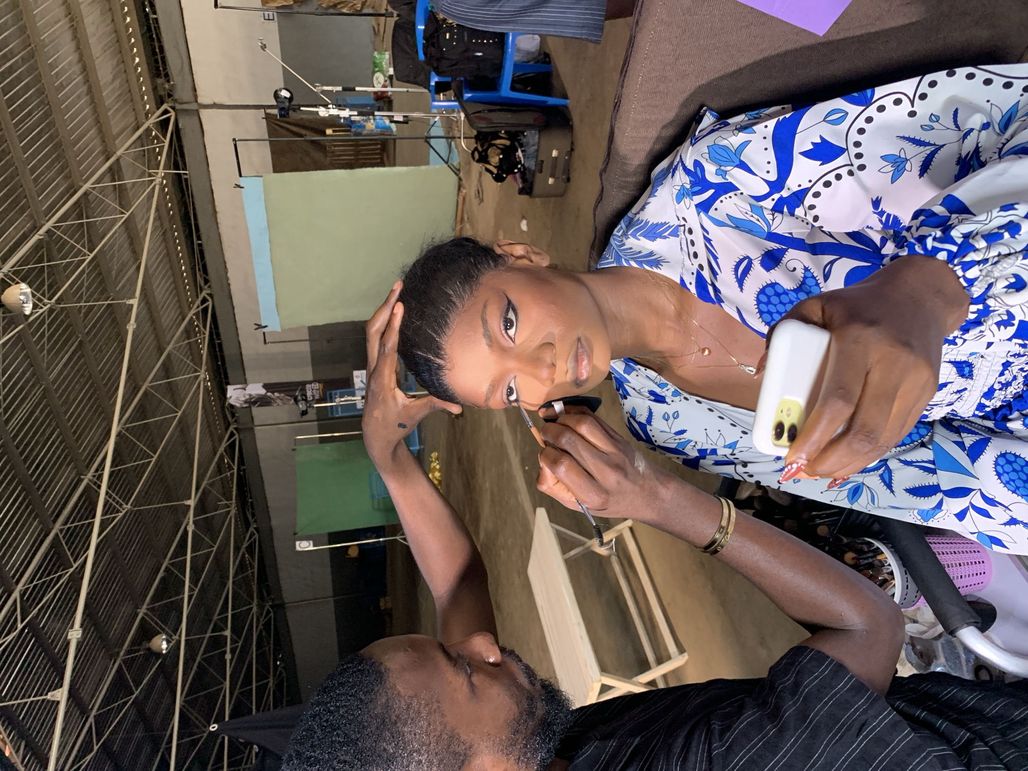 The image depicts a behind-the-scenes moment in what appears to be a photography or film studio located in Barakin Lerawi, Nigeria. A male makeup artist, wearing a black pinstriped top, is meticulously applying eyeliner to a female model. The model, seated and wearing a white and blue patterned top, holds a white smartphone in her right hand. The environment is an indoor space with a high, corrugated metal roof supported by exposed beams and a visible light fixture. In the background, there are green and light blue backdrops, studio lighting equipment, chairs, luggage, and a wooden bench, suggesting a production setting. A rack of makeup brushes is visible to the lower left of the frame.