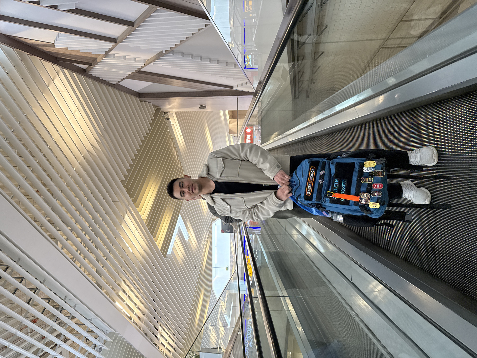 A man stands on a moving walkway, possibly at the Kazan International Airport in Kazan, Russia. He wears a beige jacket over a black shirt, black pants, and white sneakers. He has a blue backpack with multiple patches and attachments. The background consists of a modern architectural design with white slats and wooden beams. The walkway is made of metal, with a glass barrier. The lighting is bright, suggesting daylight.