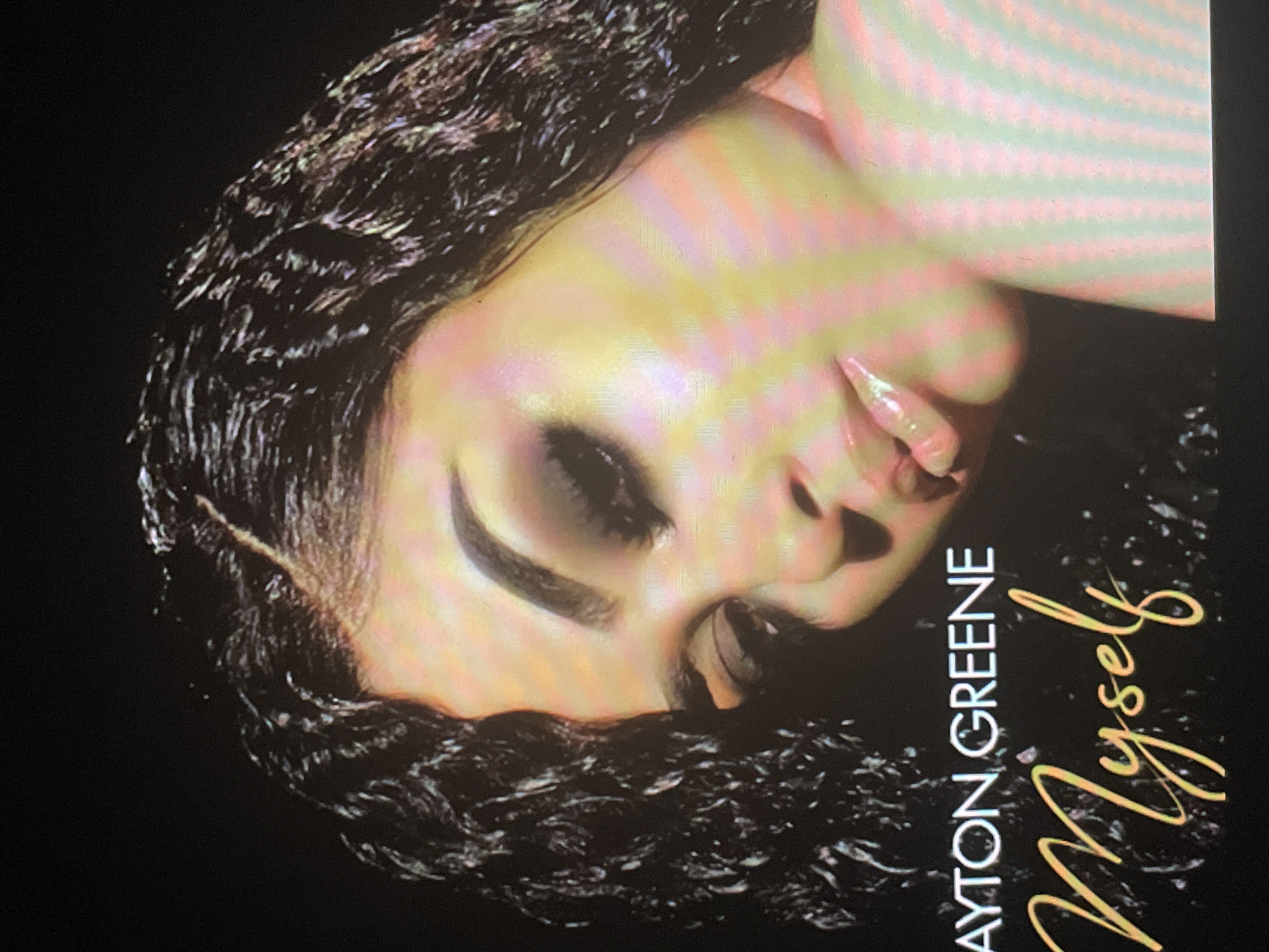 The image displays a close-up portrait of a woman, oriented as if rotated 90 degrees clockwise. Her face, primarily visible from the nose up to the forehead and left cheek, is illuminated with a soft, ethereal glow, showcasing yellow and pink/purple hues across her skin. She has dark, curly hair with a wet-look texture, framing her face. Her eyes are closed or cast downwards, accented by dramatic dark eye makeup and prominent eyebrows. Her lips are slightly parted and appear glossy.
The background is solid black, drawing attention to the subject. The overall impression is serene and artistic, suggesting a contemplative or pensive mood.
Text is visible on the right side of the image (when oriented correctly): "DAYTON GREENE" is written vertically in an all-caps, sans-serif font, and "Myself" is written horizontally in a flowing, cursive script next to it. Both text elements are in a light yellow or gold color. The image quality suggests it may be a photograph of a digital screen, with subtle scan lines and color artifacts visible.