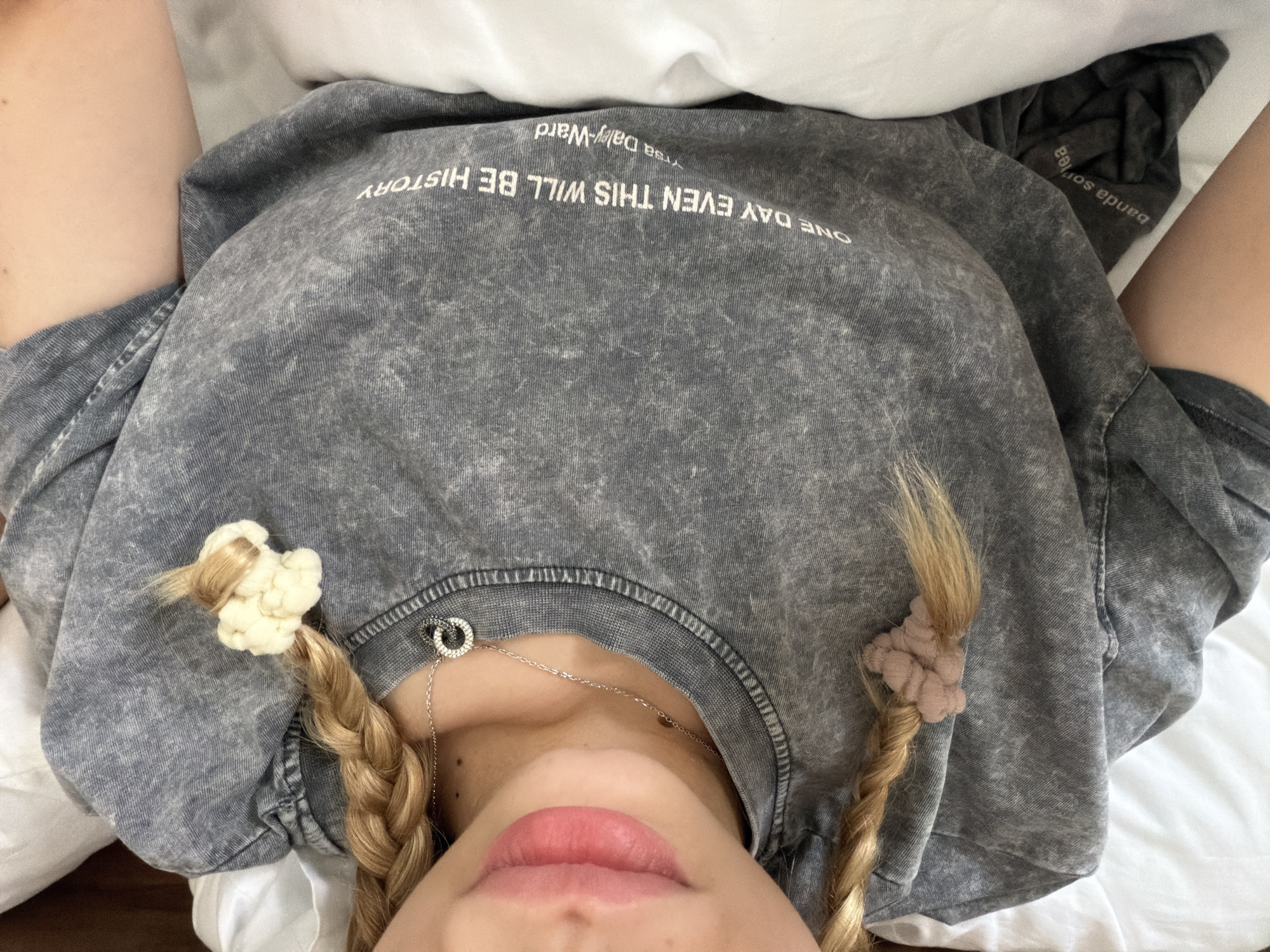 A view looking down at a person lying on a white surface, likely a bed. The person wears a gray acid-wash t-shirt with the printed words "ONE DAY EVEN THIS WILL BE HISTORY" and "ytsa Daly-Ward." The person has blonde braided hair with a white floral hair tie on one side and a tan-colored scrunchie on the other. A silver necklace with a circular pendant is visible around their neck. Their lips are visible at the bottom of the frame, painted with pink lipstick. The location is not discernable from the image; Đà Nẵng, Vietnam cannot be determined.
