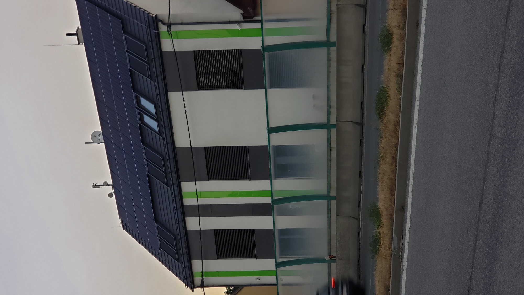 The image shows a modern building with solar panels on the roof, located near a road in Prešov, Slovakia. The building has a distinctive facade with vertical stripes of white, dark gray, and bright green. There are several windows with dark blinds on the upper floor. A satellite dish and antennas are visible on the roof. A transparent barrier, possibly for noise reduction, separates the building from the road. A blurred vehicle is seen driving on the road in the foreground, indicating movement. The sky is overcast and the overall lighting suggests it is either early morning or late evening. The vegetation along the roadside is dry and yellowed.
